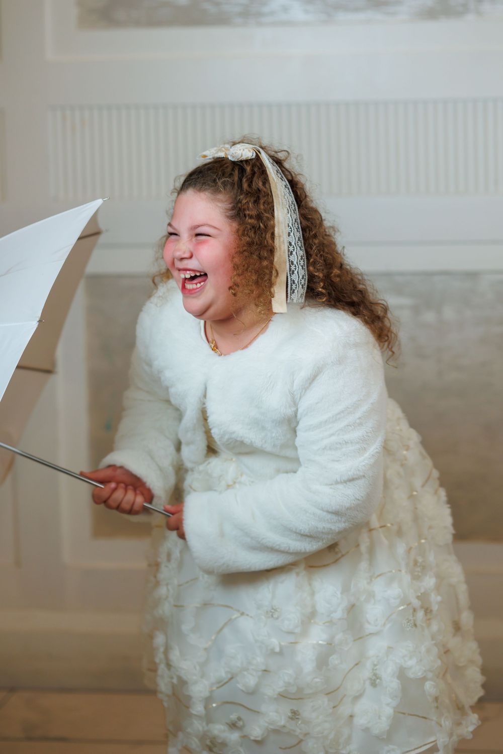 A joyful girl in a white fluffy dress and bow laughs while holding a white umbrella on elegant stairs.