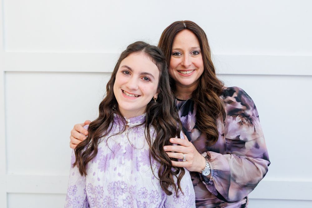 Two smiling women pose together, wearing matching purple and gray tie-dye outfits against a white background.
