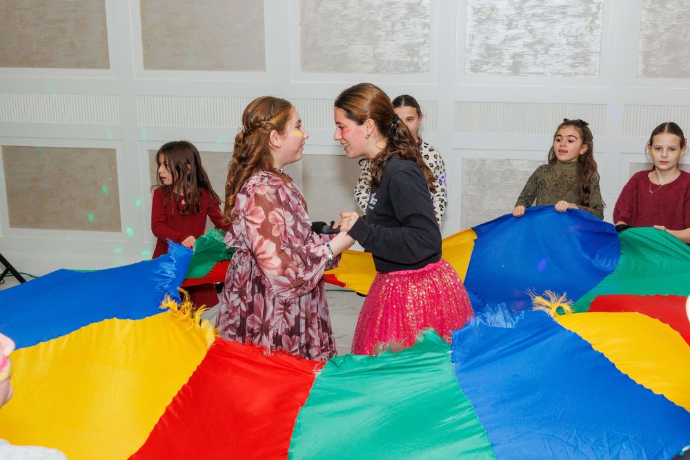 Children playing with a colorful parachute indoors at a party or school event.