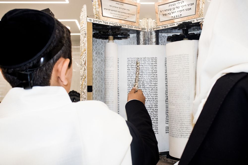 Two Jewish men in traditional white prayer shawls reading from a Torah scroll during a religious ceremony.