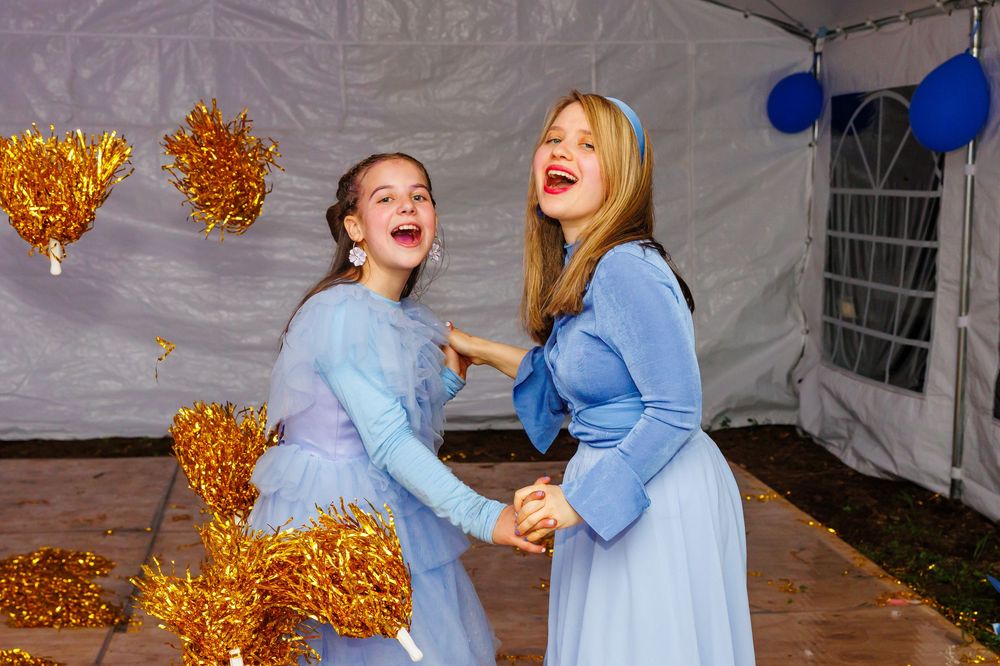 Two girls in matching light blue dresses laugh and dance together at a festive event with gold pom-poms.