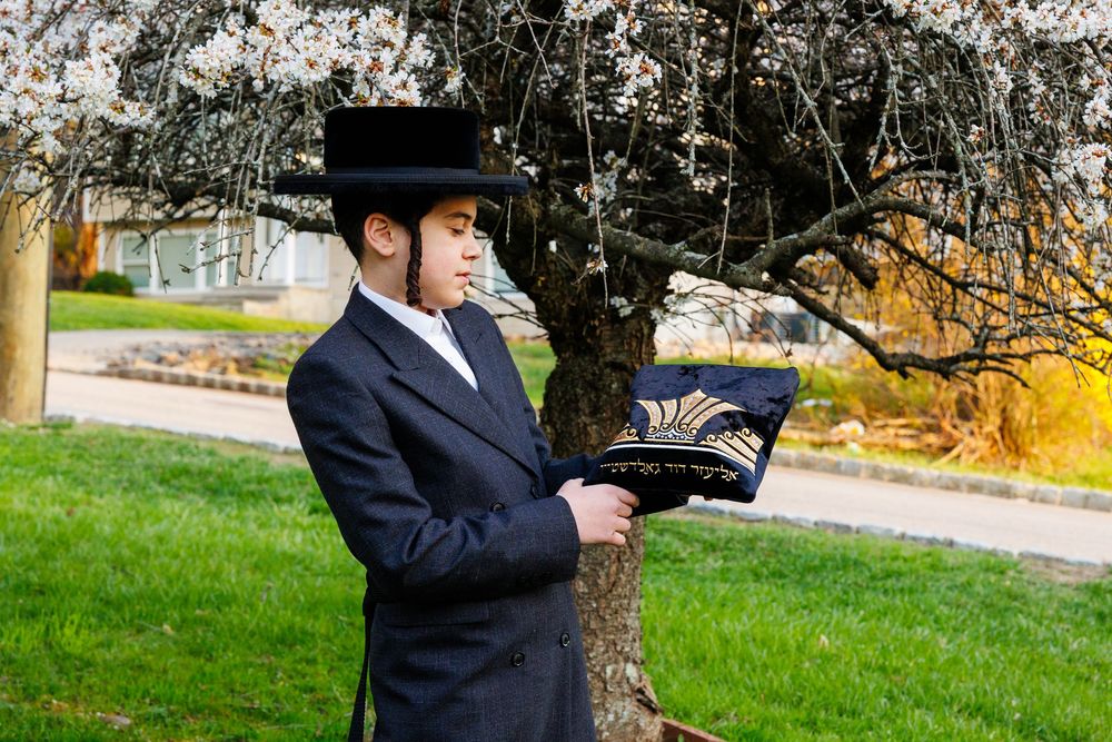 Young Orthodox Jewish man in black suit and hat holding a decorative prayer book bag under a blossoming tree.