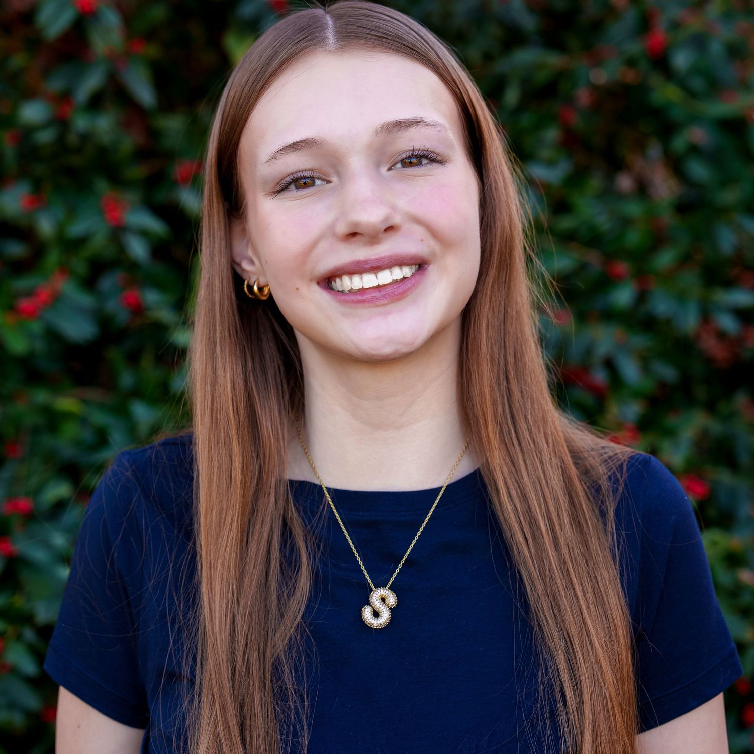 Smiling young woman with long auburn hair wearing a navy top and gold necklace outdoors.