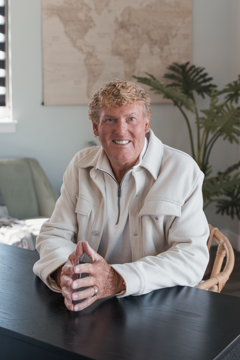 Smiling older man with curly blond hair sits at dark table in cream jacket, world map on wall behind him.