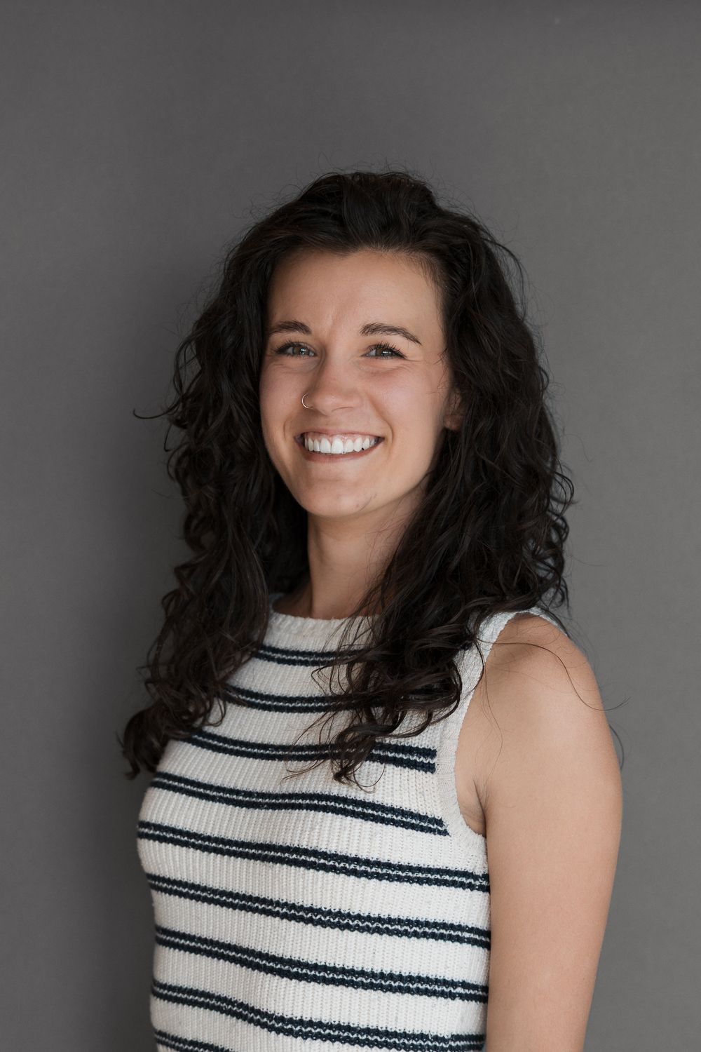 Smiling young woman with curly dark hair wearing a striped sleeveless top against a gray background.