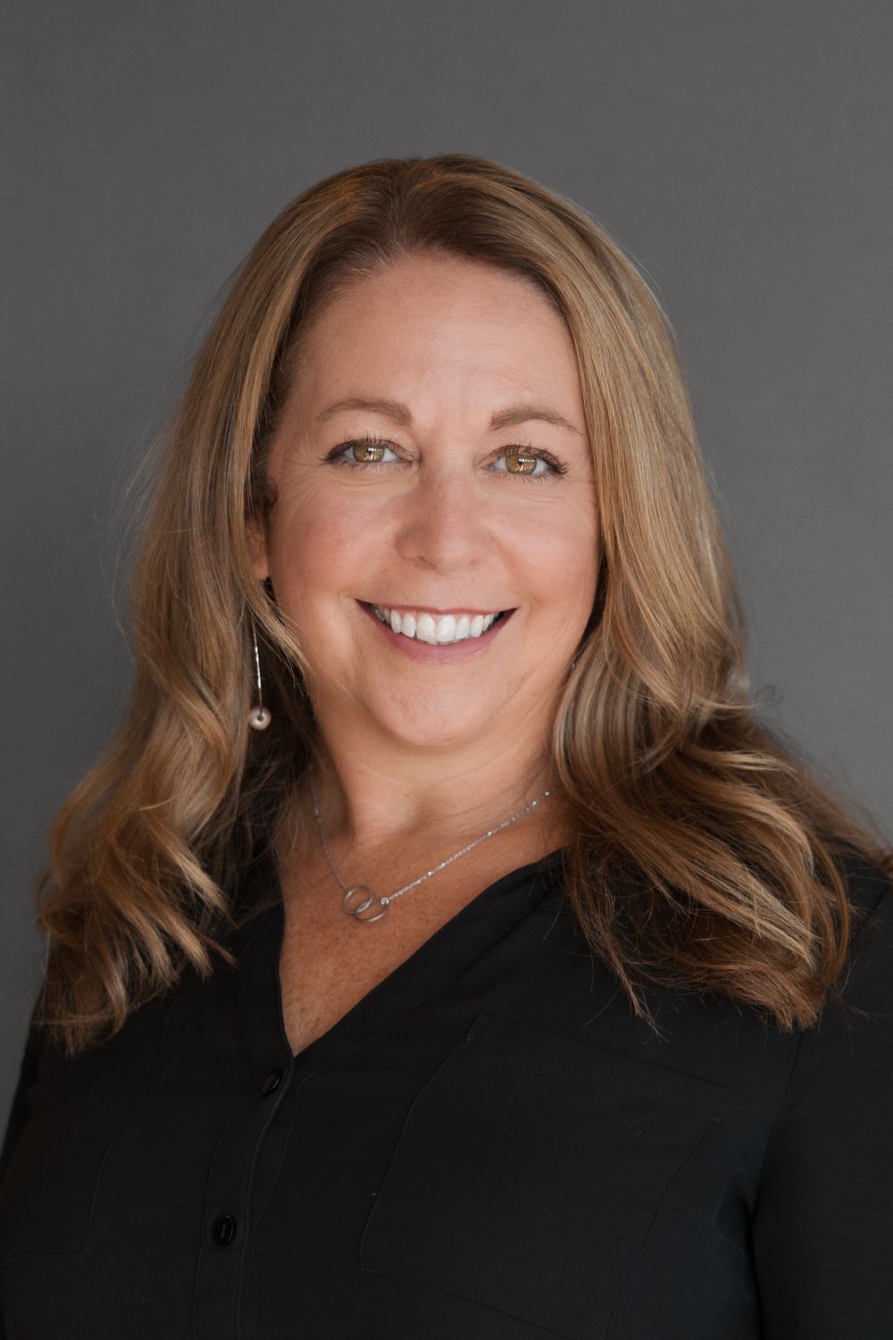 Professional headshot of a smiling woman with blonde hair wearing a black top and delicate necklace on gray background.