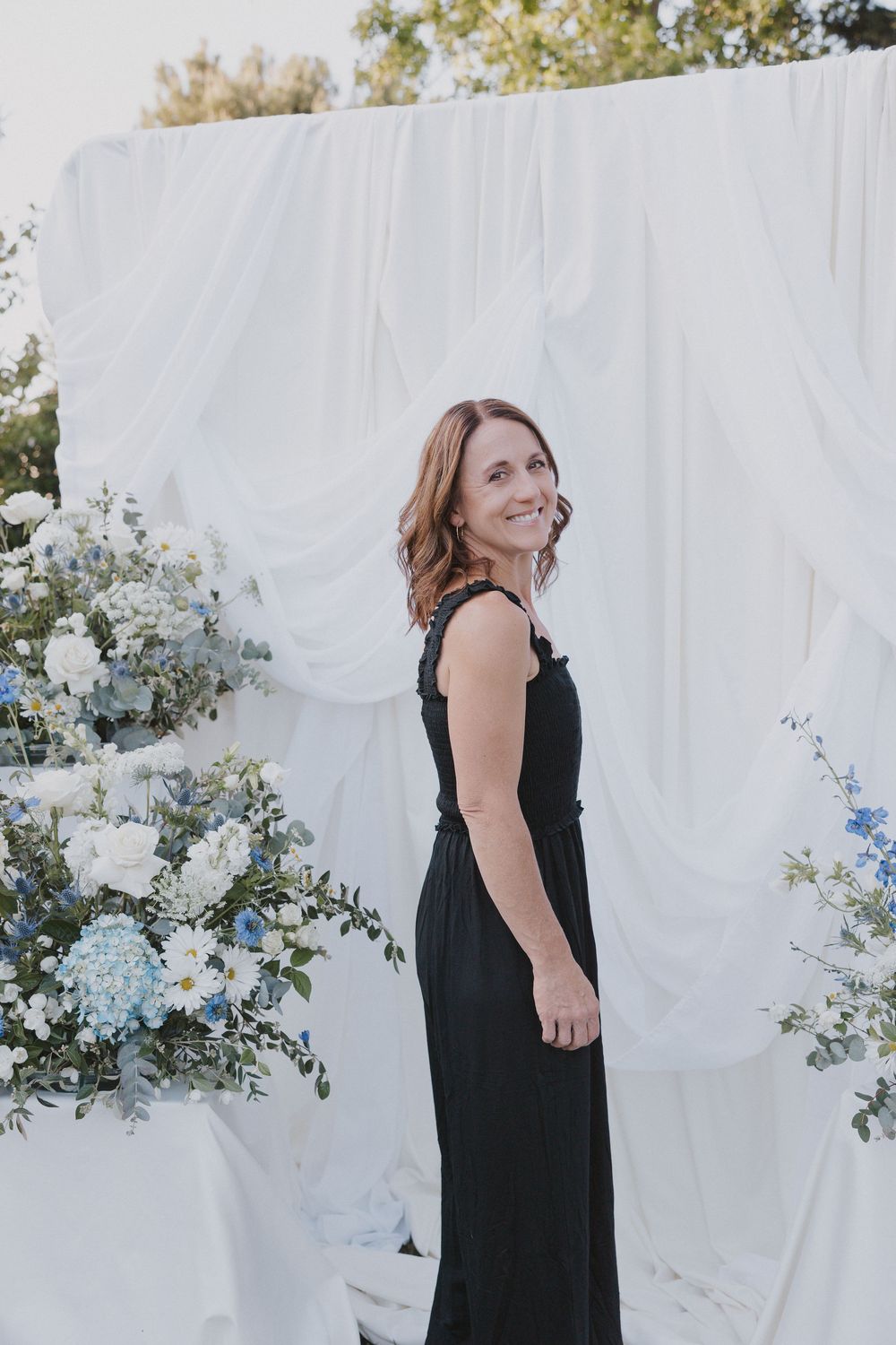 Woman in black dress smiling at outdoor wedding ceremony with white draping and blue floral arrangements.