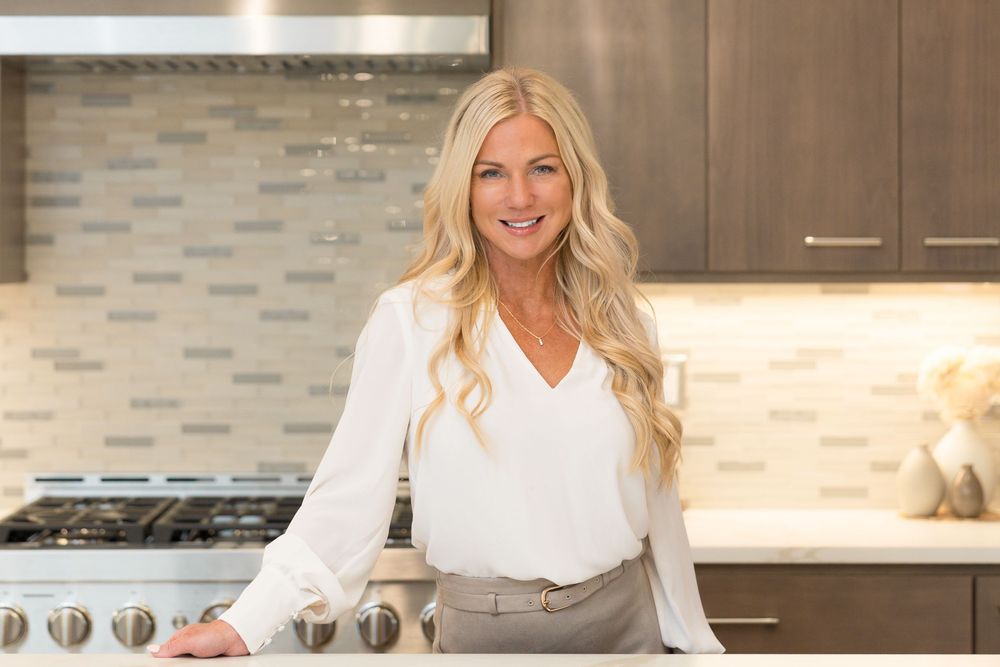 Smiling blonde woman in white blouse leaning on kitchen counter with modern backsplash and stainless stove.
