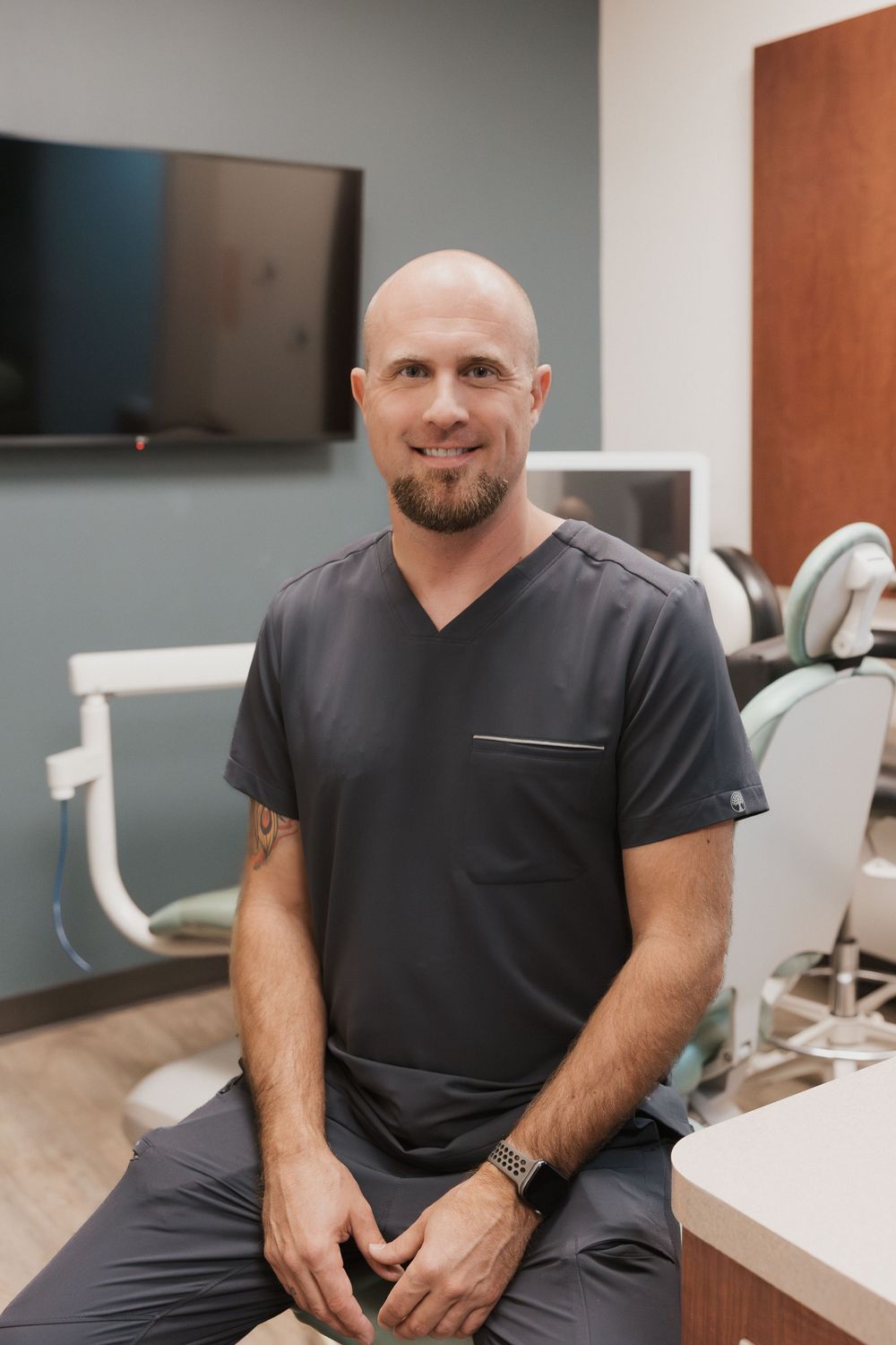 Smiling bald male dentist in gray scrubs sitting in a modern dental office with equipment visible.