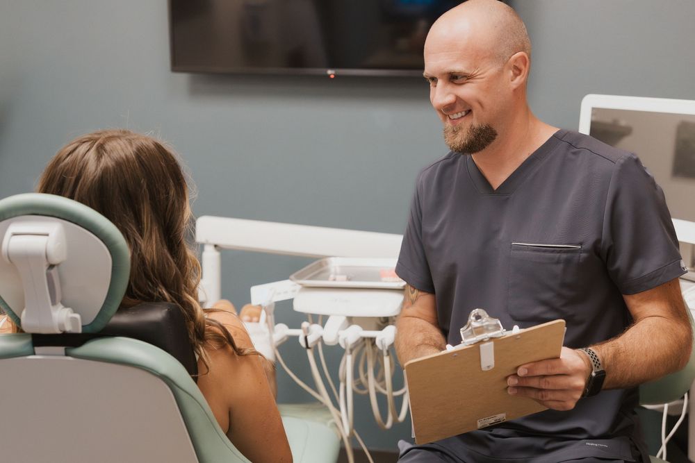Dentist in scrubs smiling while reviewing clipboard with patient seated in dental chair during consultation.