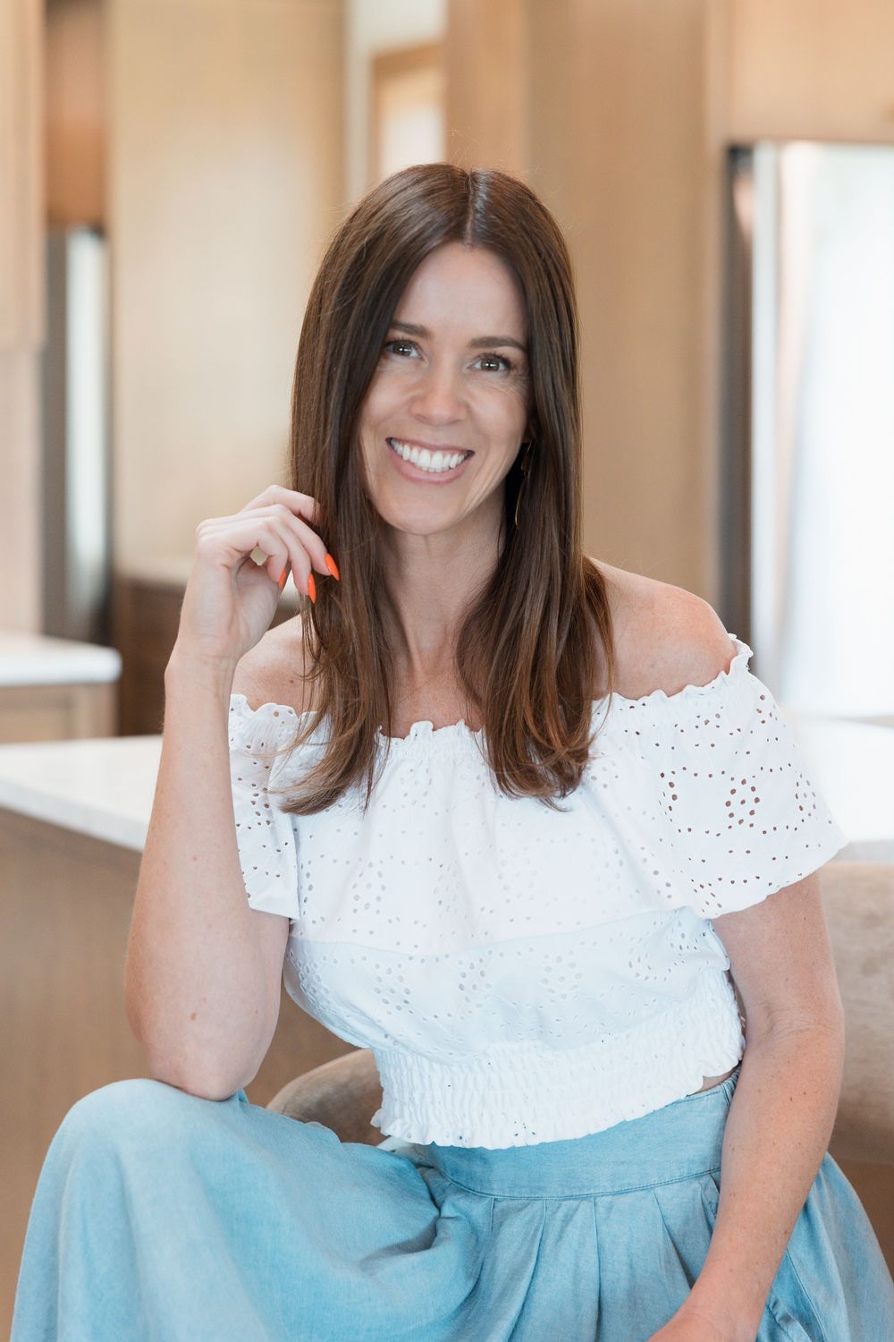 Smiling brunette woman in white off-shoulder eyelet top and light blue pants seated indoors.