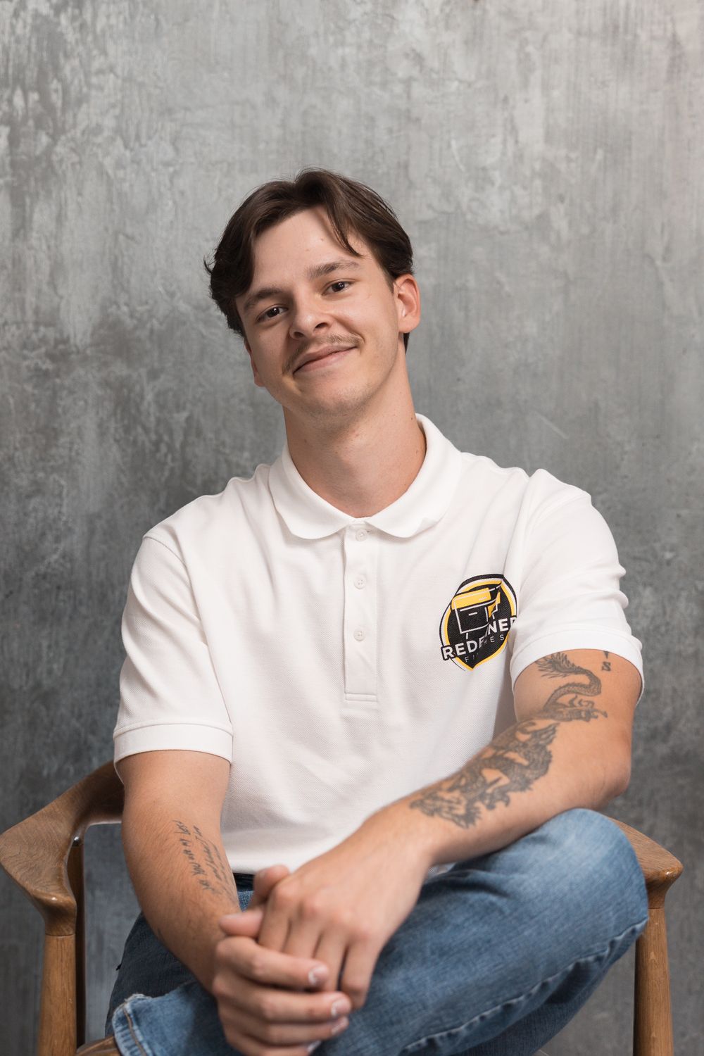 Young man with tattoos smiling in white polo shirt sitting relaxed on wooden chair against gray concrete wall.