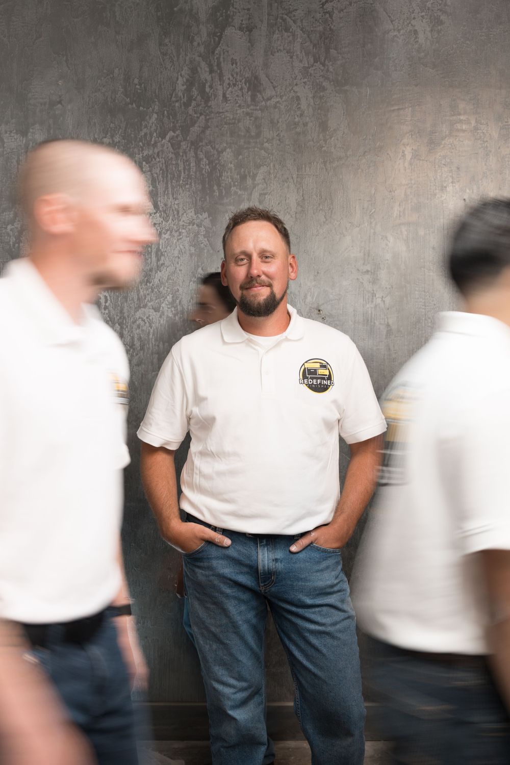 Man in white polo shirt stands confidently against gray wall while colleagues walk past in motion blur.