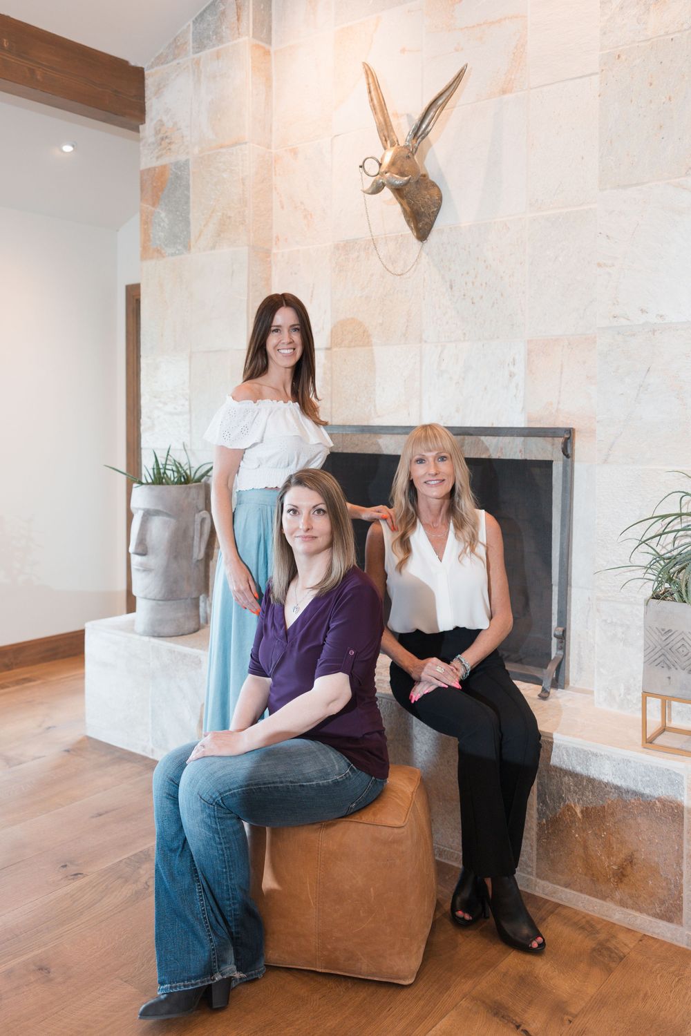 Three women posing elegantly near a stone fireplace in a modern interior with wood floors.
