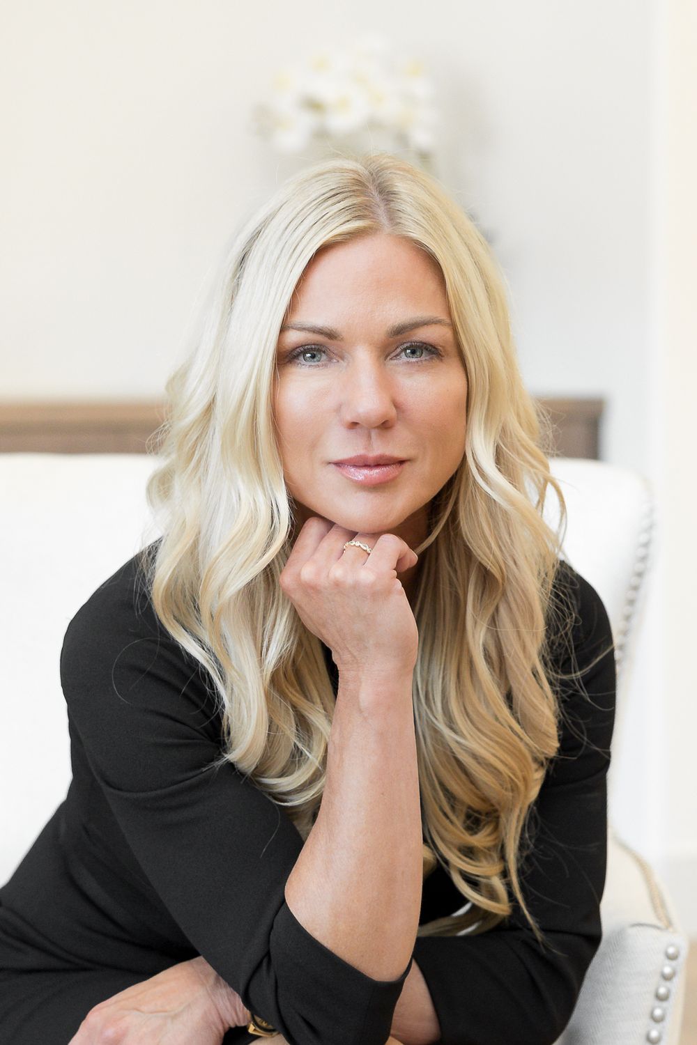 Blonde woman in black top resting chin on hand, seated in white chair with elegant interior background.