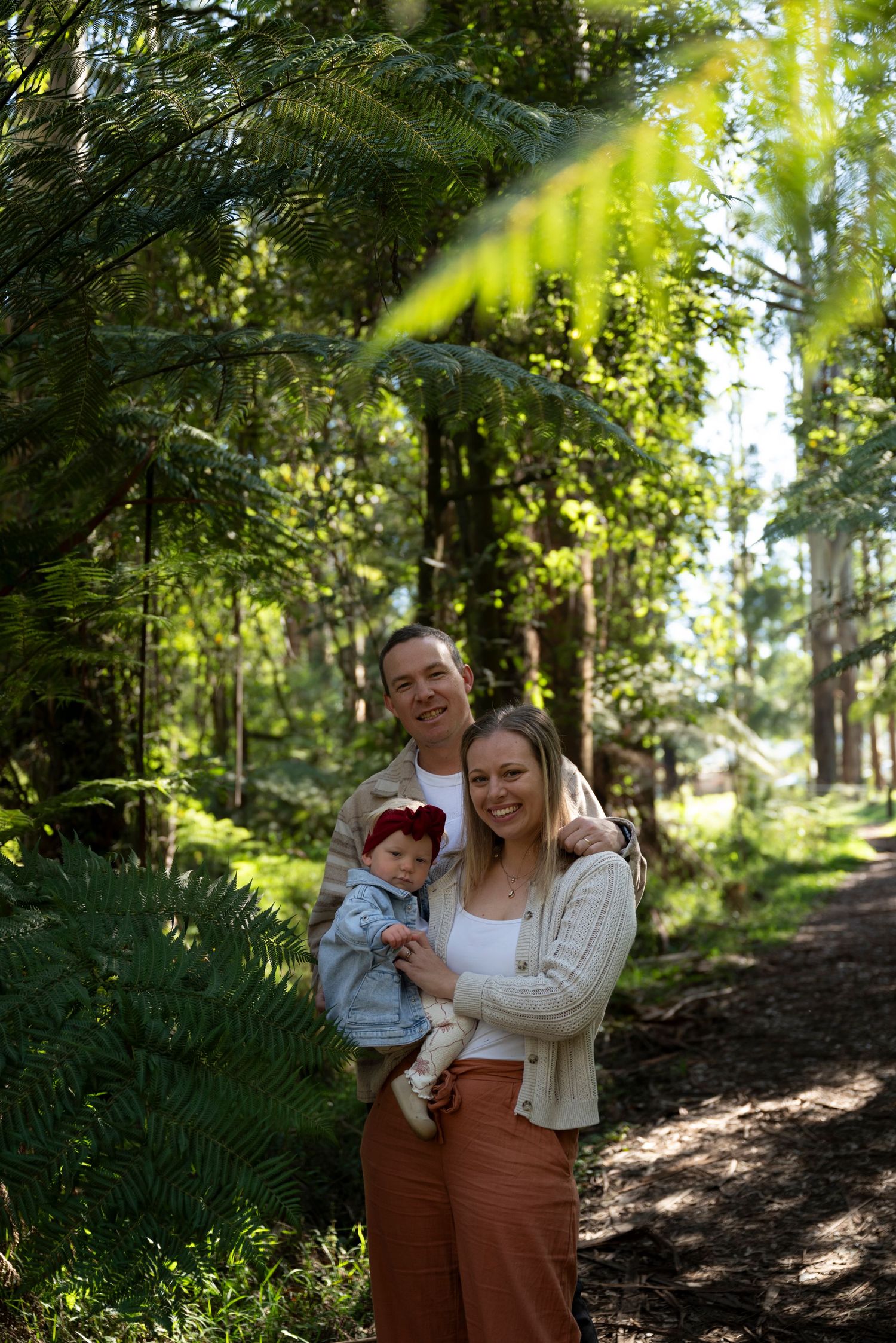 A happy young family with a baby poses on a sunlit forest path surrounded by lush green ferns and tall trees.