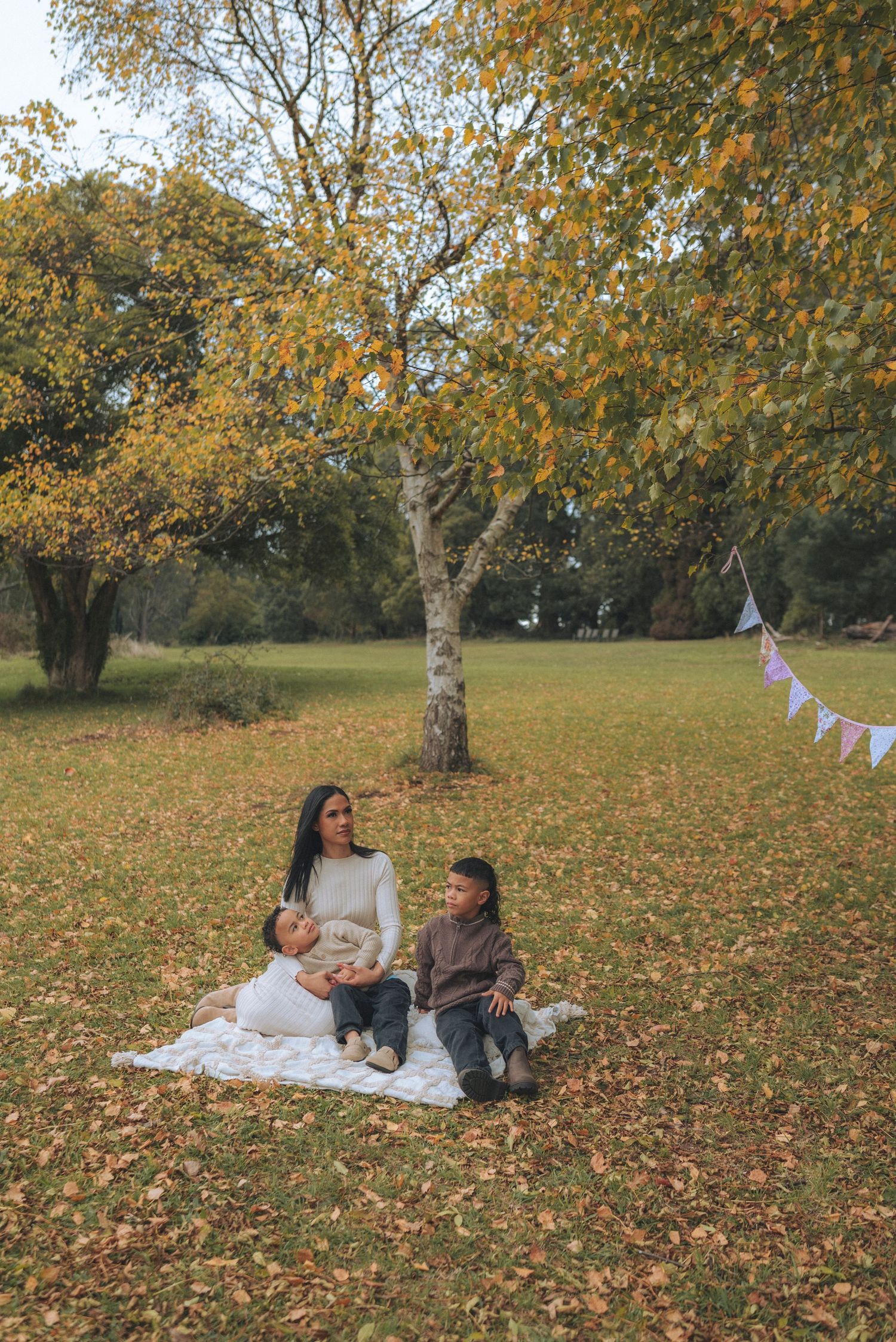 A mother sits on a blanket with her two children in an autumn park, surrounded by golden leaves and trees.