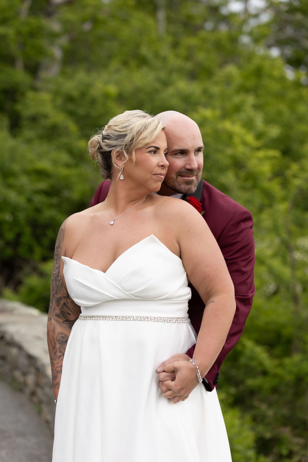 Bride in white gown and groom in burgundy suit pose outdoors with lush green trees in background at wedding.