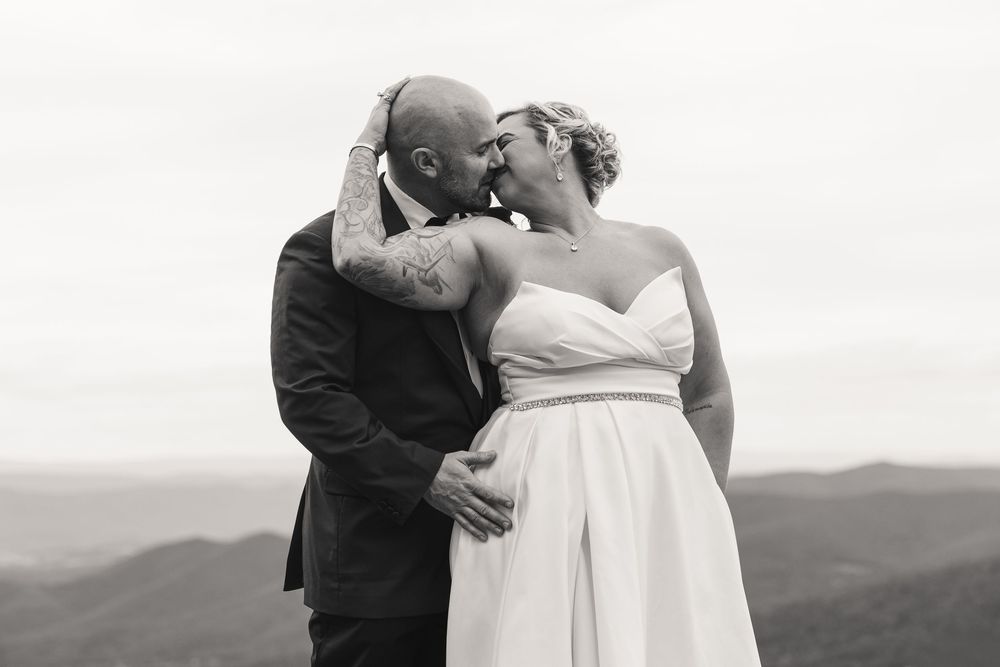 Black and white photo of a tattooed groom kissing his bride in a white dress with mountain views behind them.