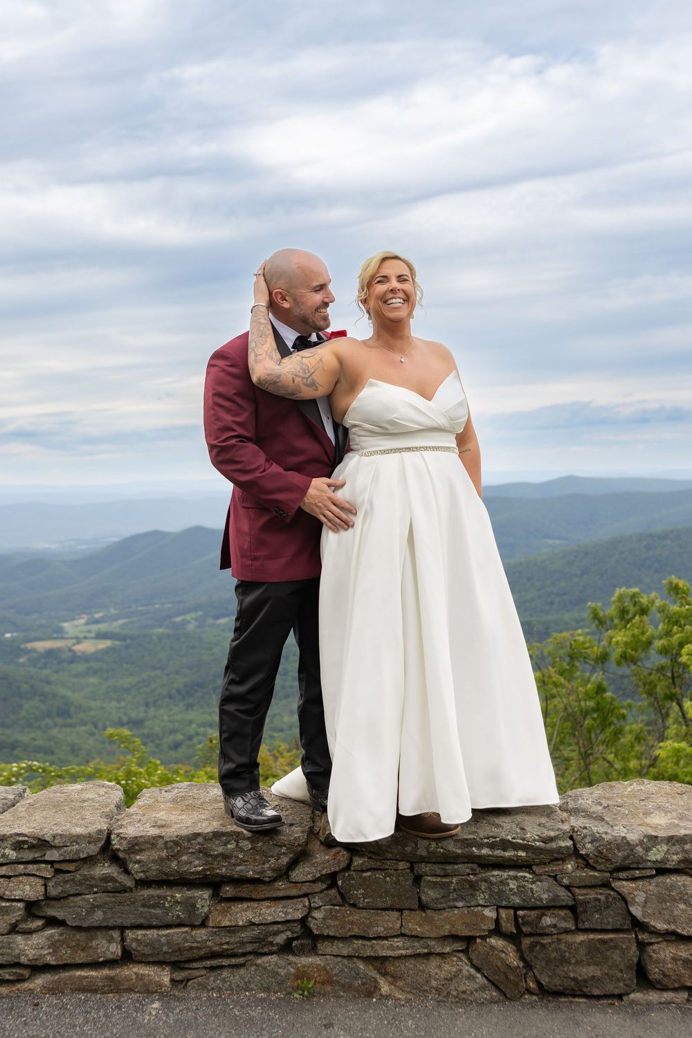 Newlywed couple posing on stone wall with mountain views, bride in white gown and groom in burgundy suit.