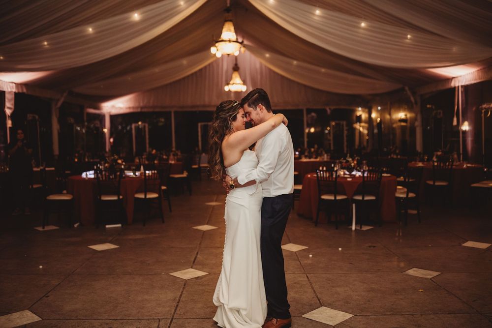 Bride and groom share first dance in elegant tented wedding reception with warm amber lighting and draped ceiling.