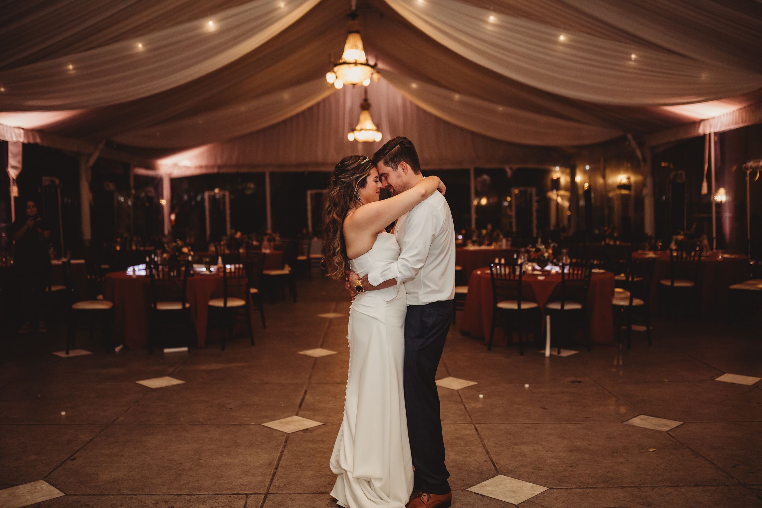 Bride and groom share first dance in elegant tented wedding reception with warm amber lighting and draped ceiling.