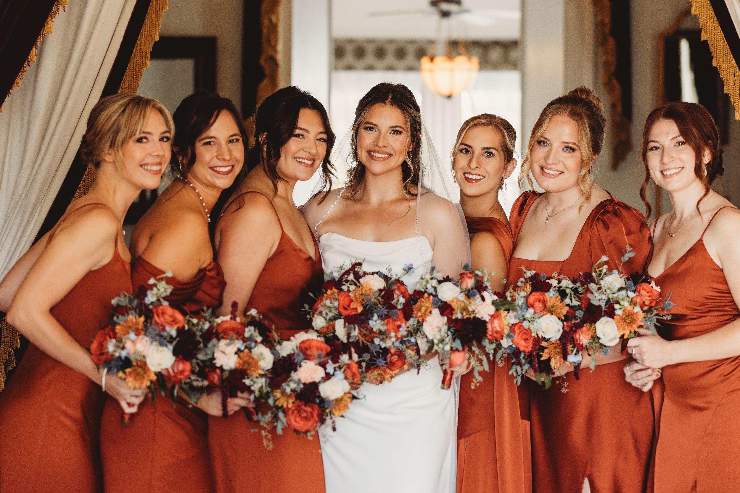 Bride in white gown with six bridesmaids in rust orange dresses holding floral bouquets at an elegant wedding venue.