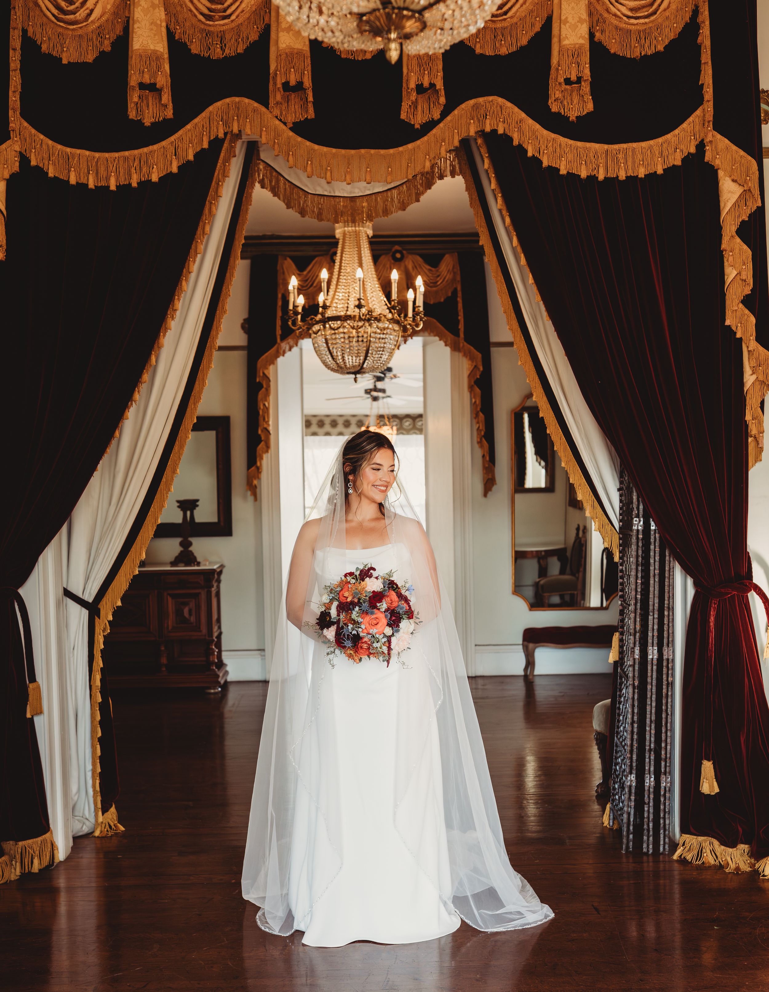 Bride in white gown and veil holds colorful bouquet under chandelier framed by gold-trimmed curtains in elegant venue.