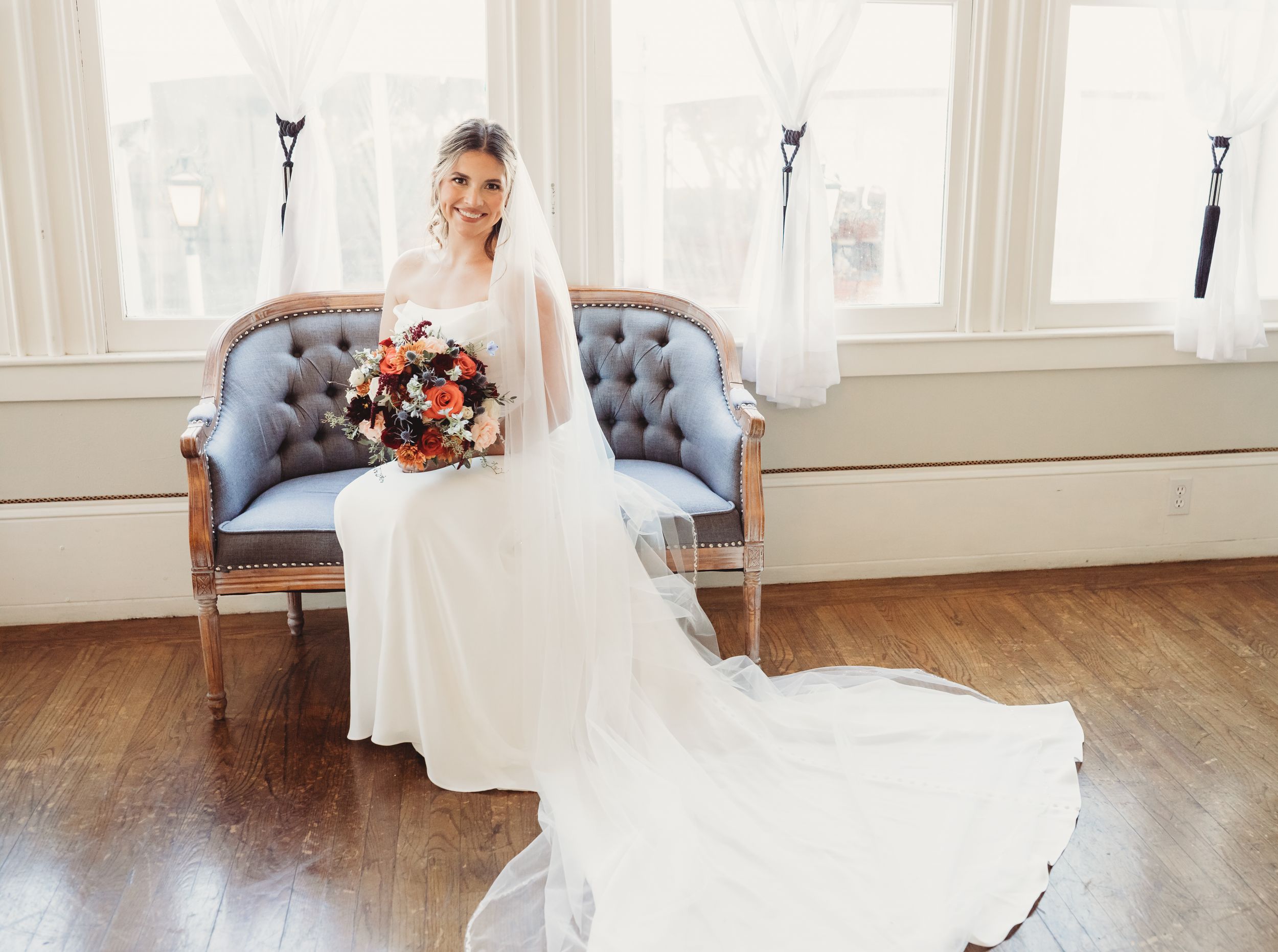 Bride in white gown with veil sits on vintage blue sofa holding colorful bouquet by bright windows on hardwood floor.