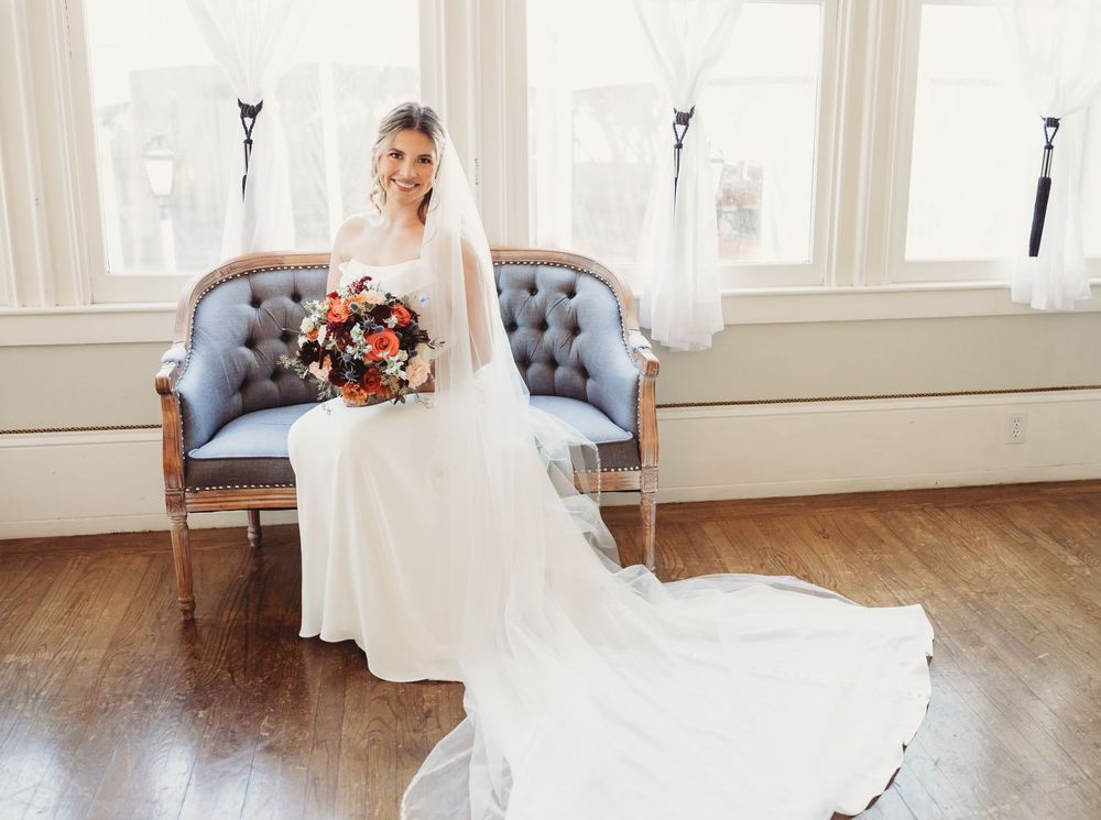 Bride in white gown with veil sits on vintage blue sofa holding colorful bouquet by bright windows on hardwood floor.