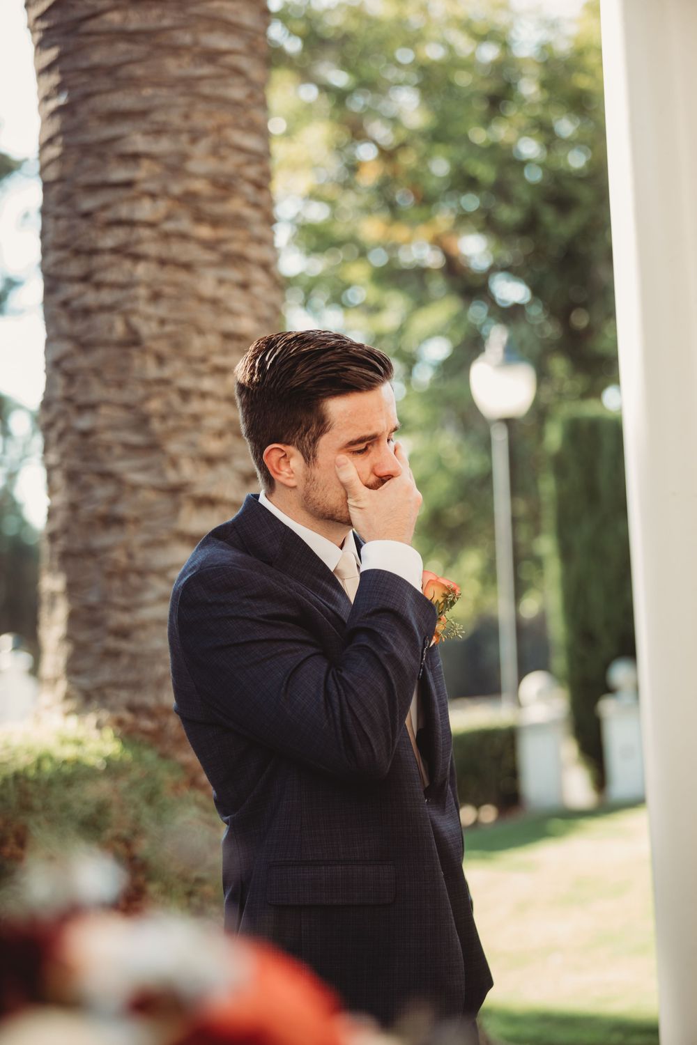 Emotional groom in navy suit with boutonniere covering mouth during outdoor wedding ceremony near palm tree.
