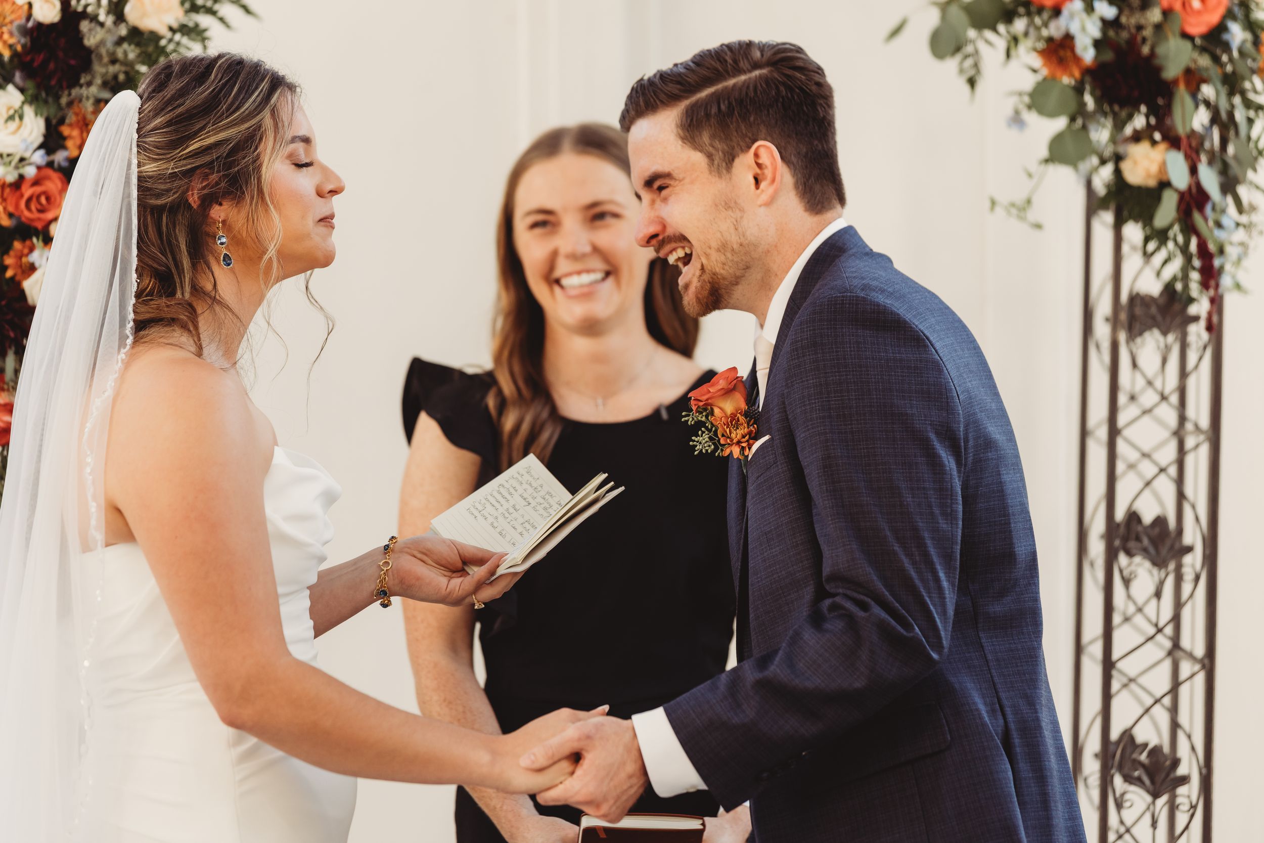 Bride and groom laughing with officiant during joyful wedding ceremony with floral arch backdrop.