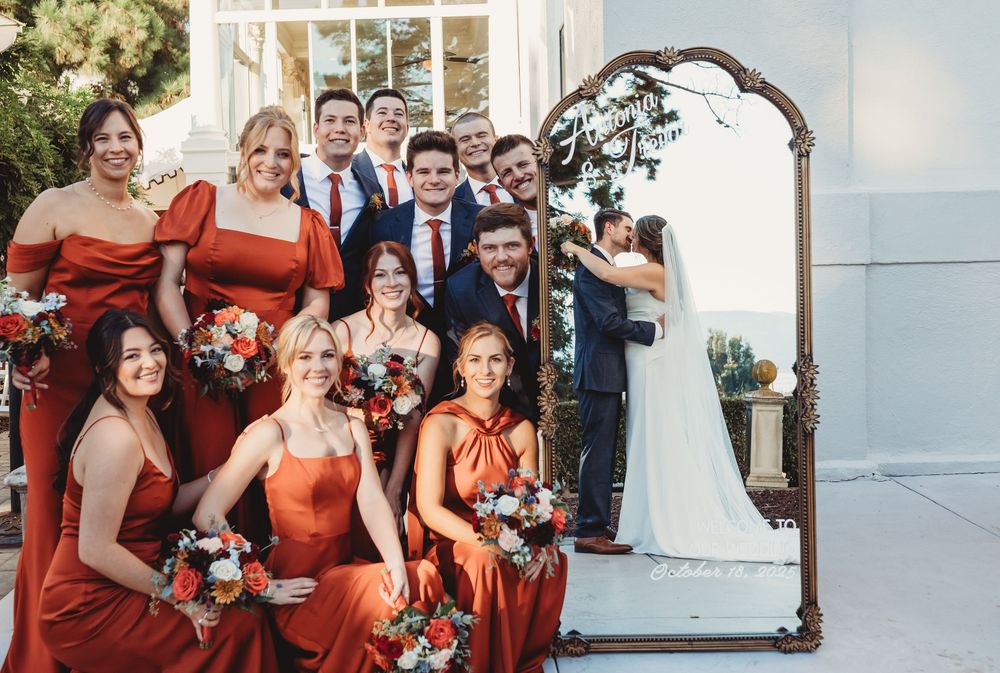 Wedding party in rust-orange bridesmaid dresses poses with bouquets beside a vintage mirror as bride and groom kiss.