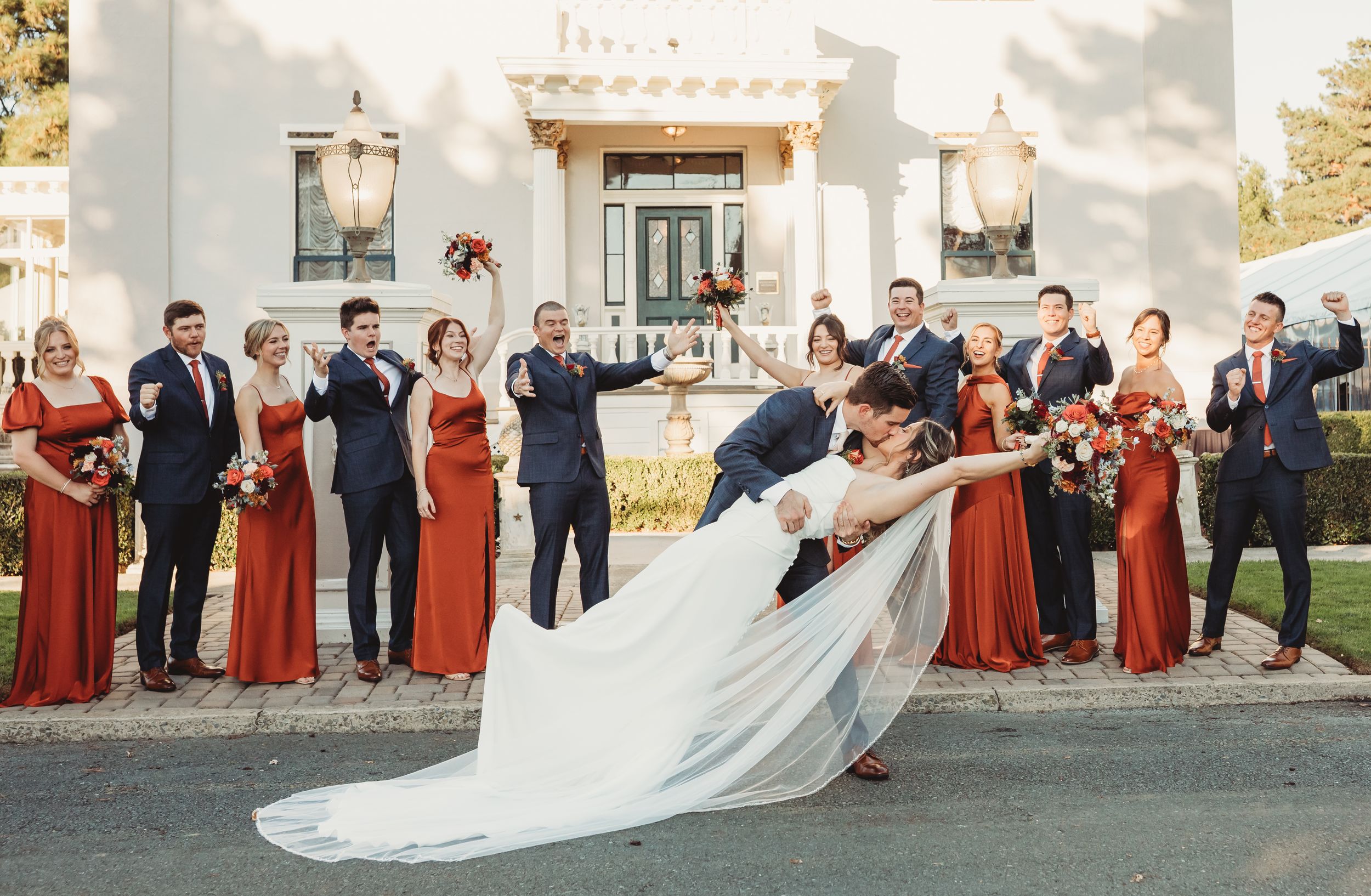 Bride and groom share romantic dip kiss surrounded by wedding party in red dresses and navy suits outside elegant white venue.