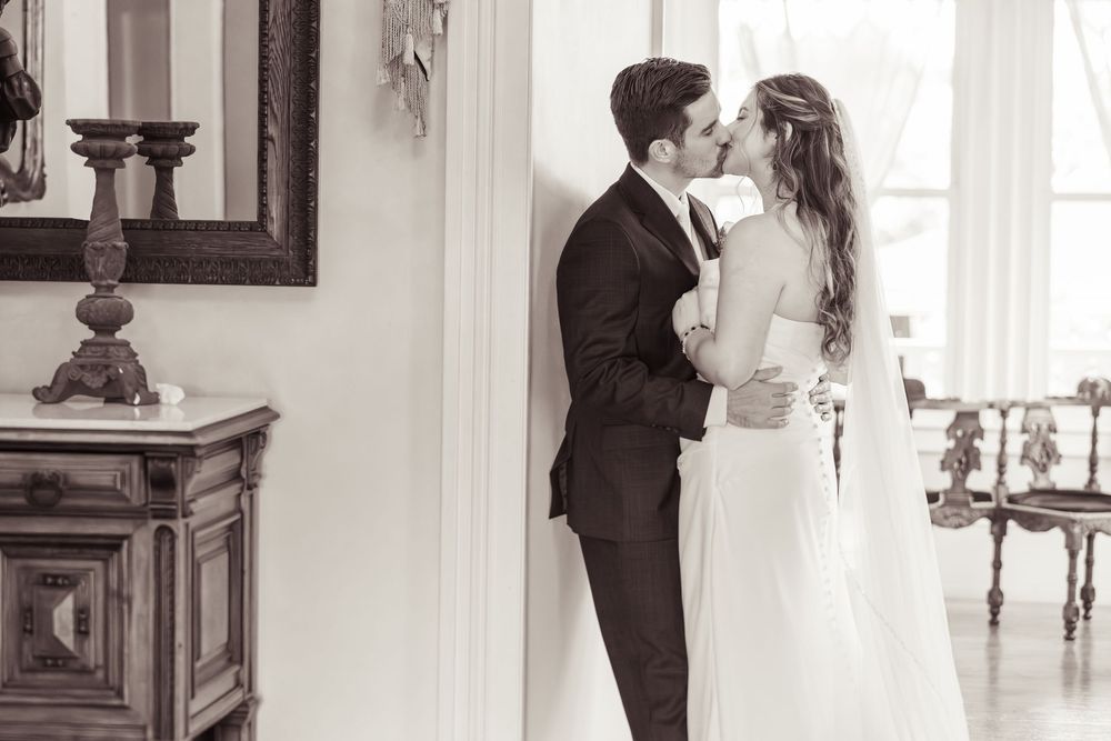 Black and white photo of a bride and groom sharing a romantic kiss indoors on their wedding day.