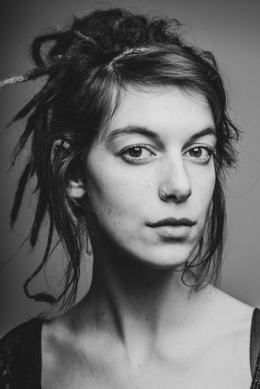 Zwart wit studioportret van een vrouw met dreadlocks en een neuspiercing, met haar hoofd licht gedraaid en haar blik in de camera.