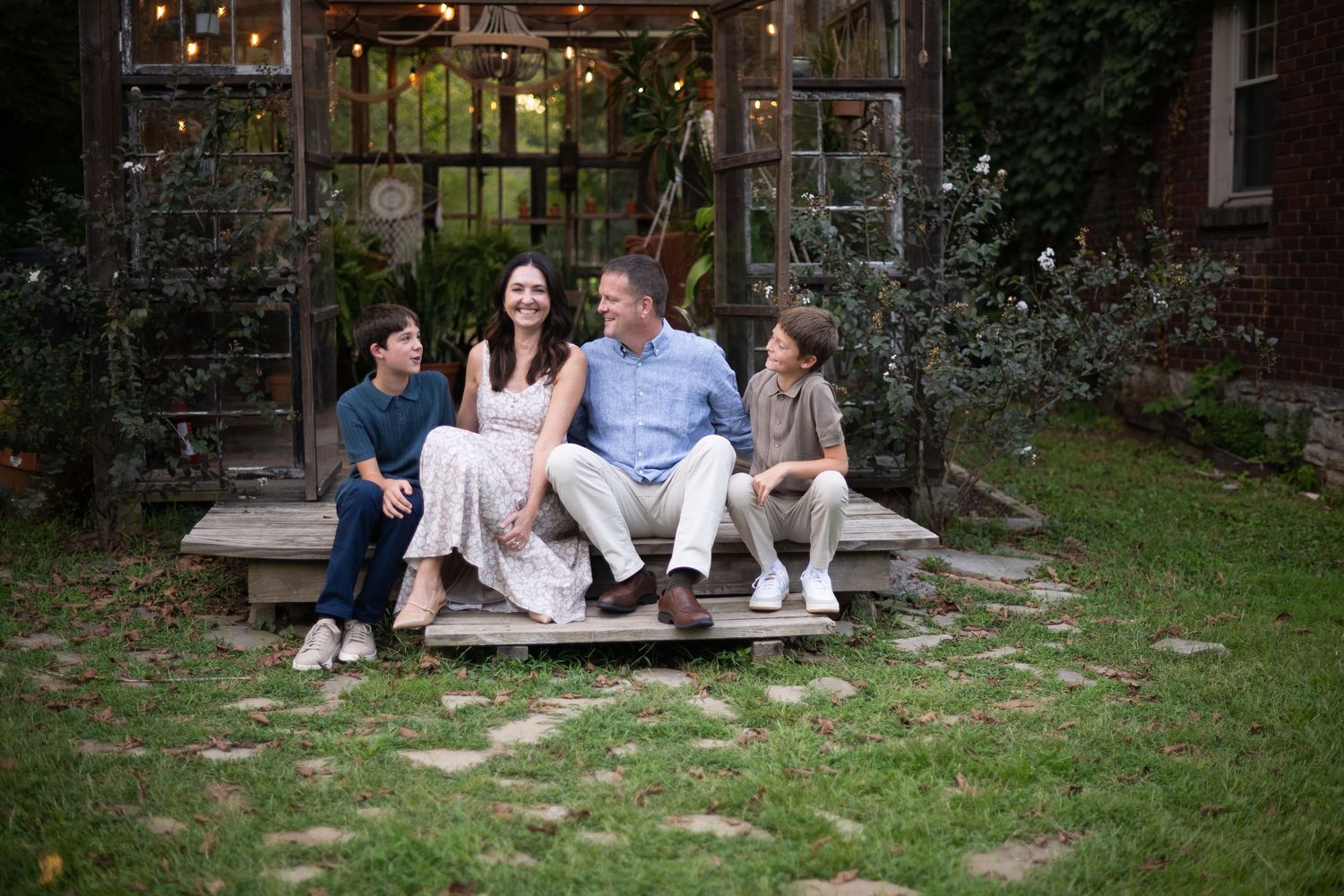 Happy family of four sitting together on wooden steps in a charming backyard garden with string lights.