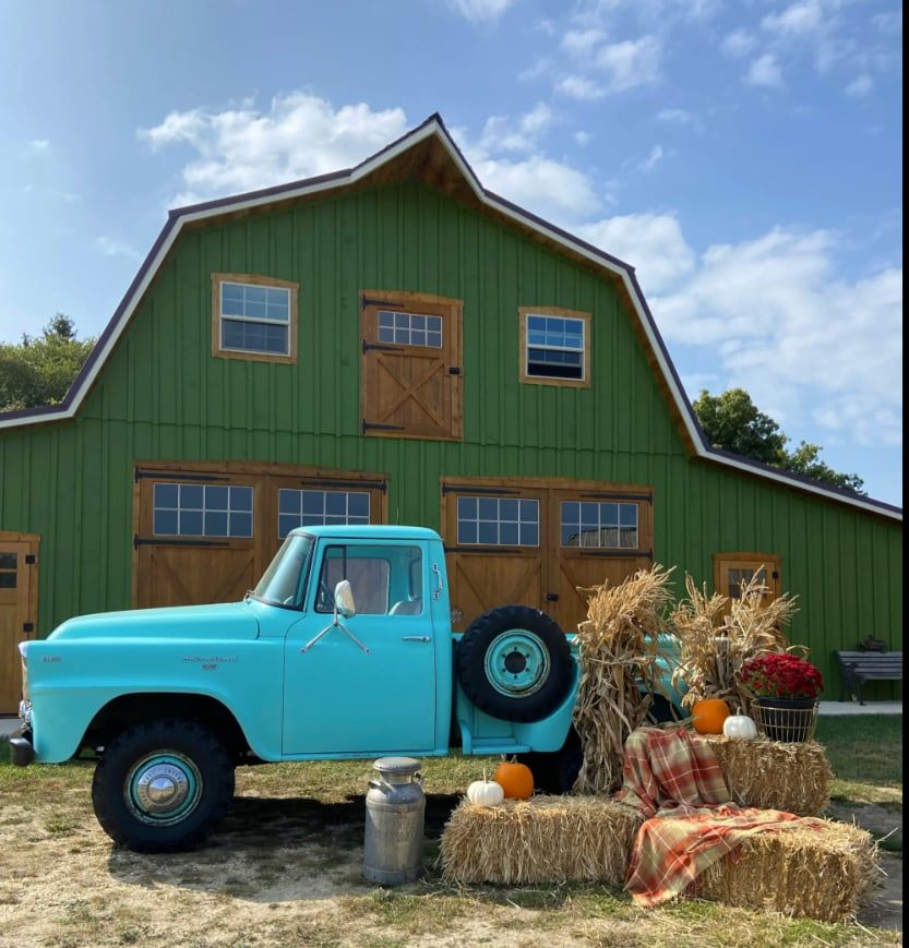 Vintage teal pickup truck parked in front of a green barn with hay bales and pumpkins in a fall farm setting.