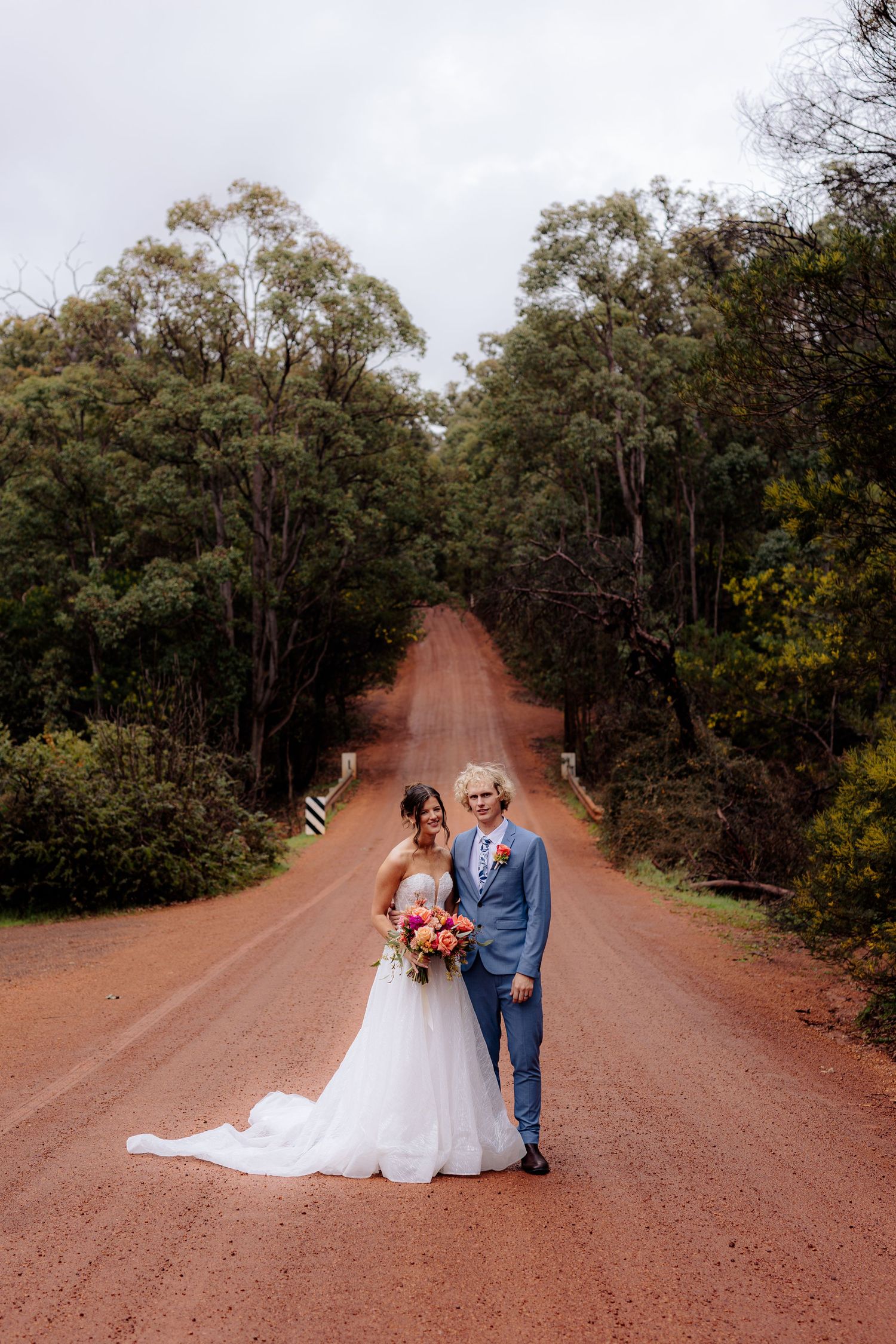 Bride and groom pose on a red dirt road surrounded by lush green trees on their wedding day.