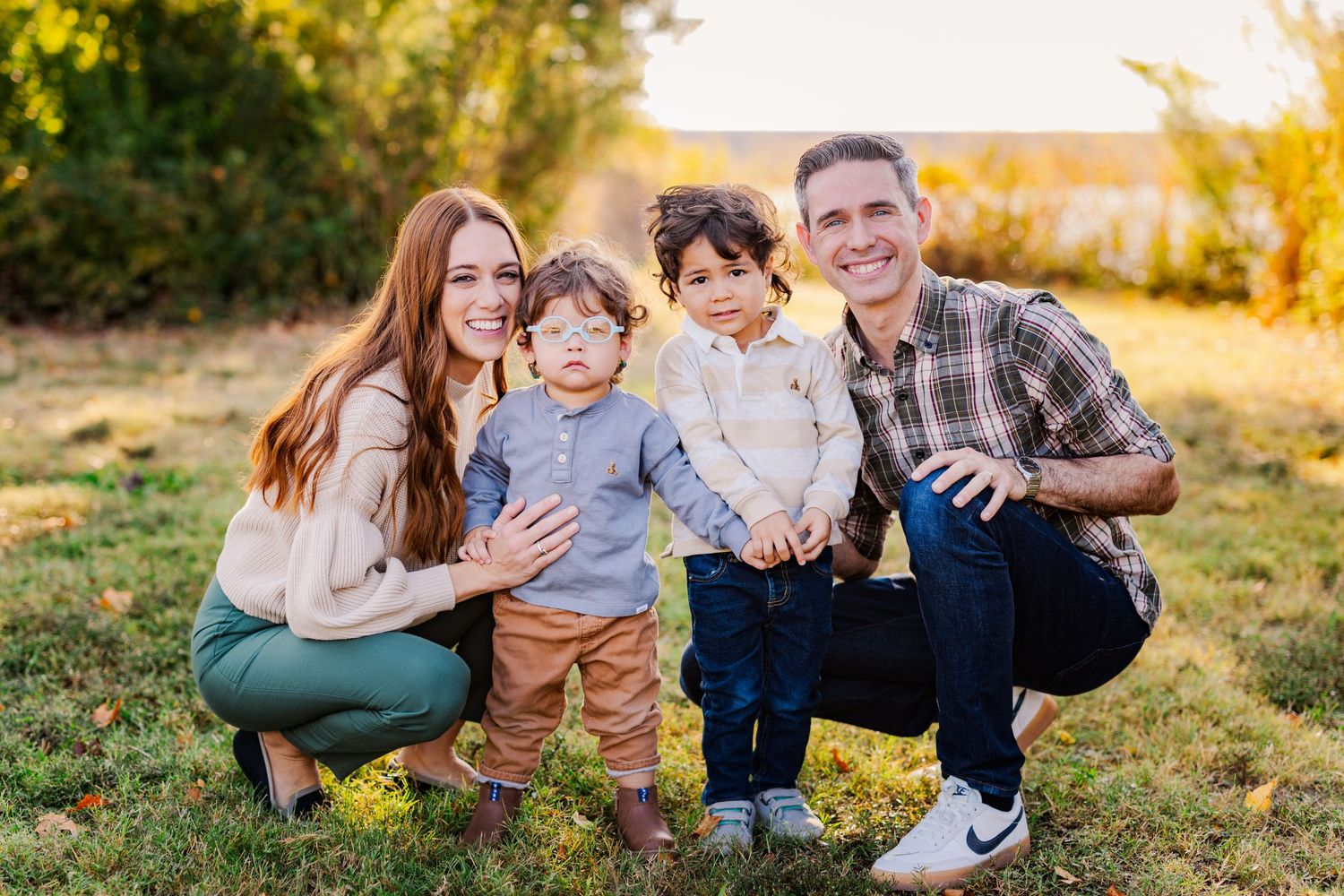 Happy family of four with two young boys smiling together in a sunny outdoor field setting.
