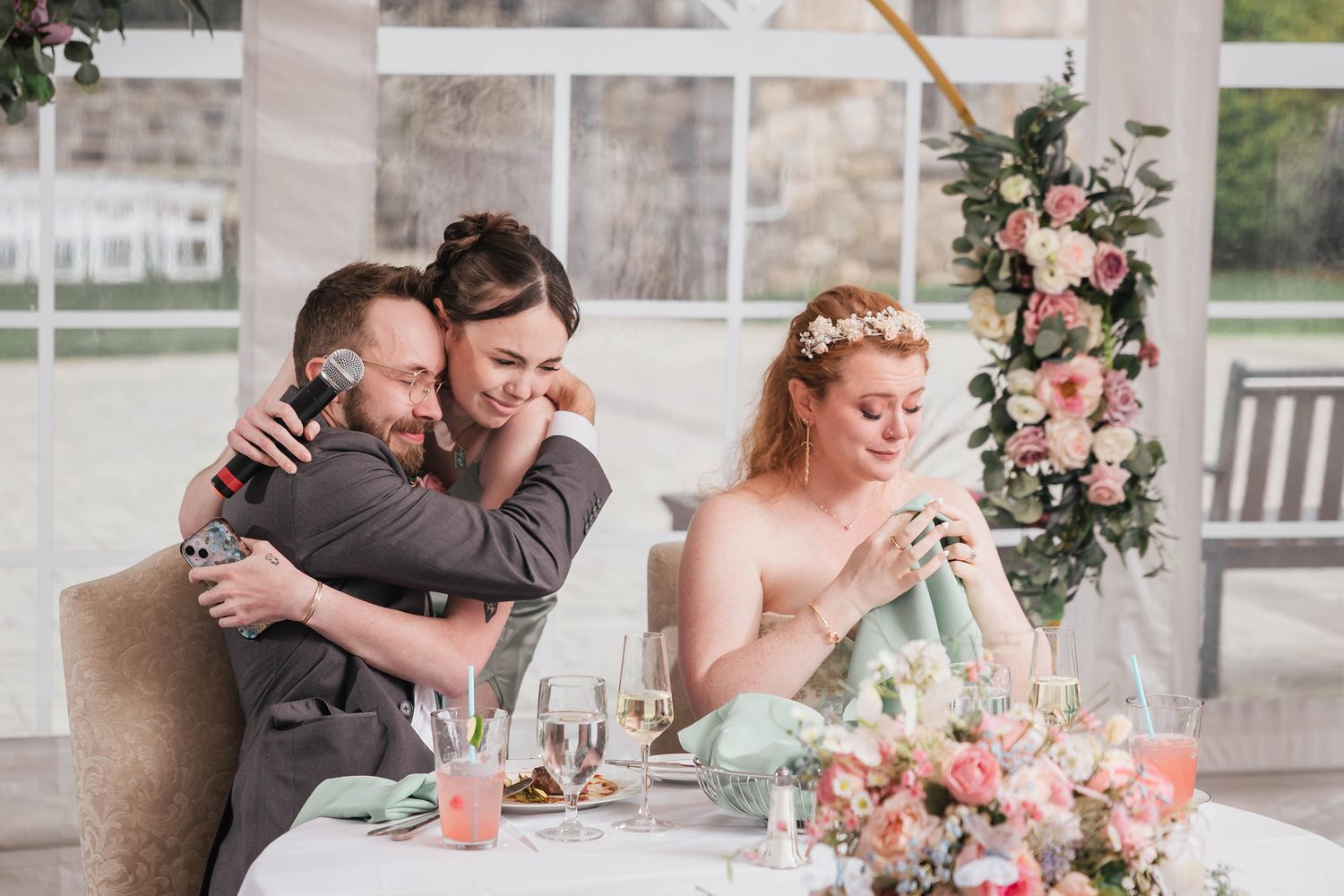 Bridesmaid hugging a groomsman tightly during a speech, bride seated beside them.