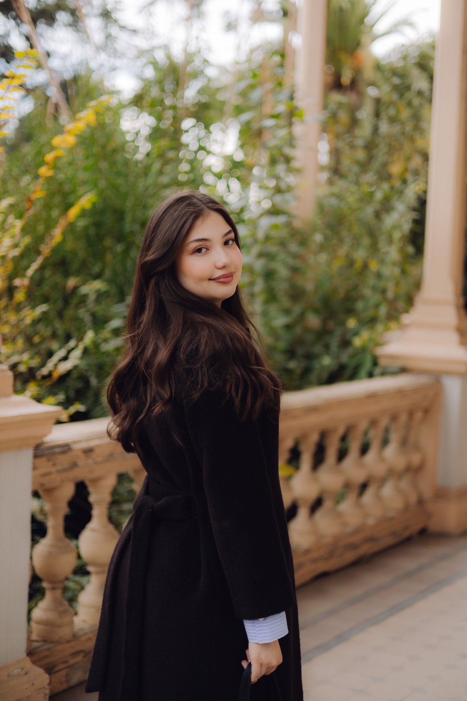 Young woman in black coat smiling over shoulder on ornate balcony with lush green garden backdrop.