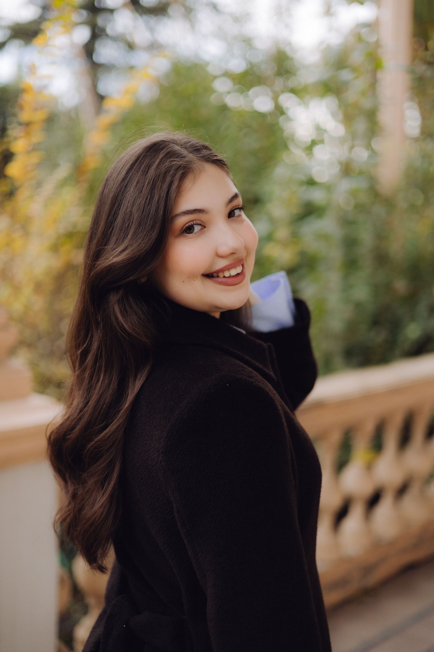 Young woman with long dark wavy hair smiling over shoulder, wearing black coat outdoors in autumn garden setting.