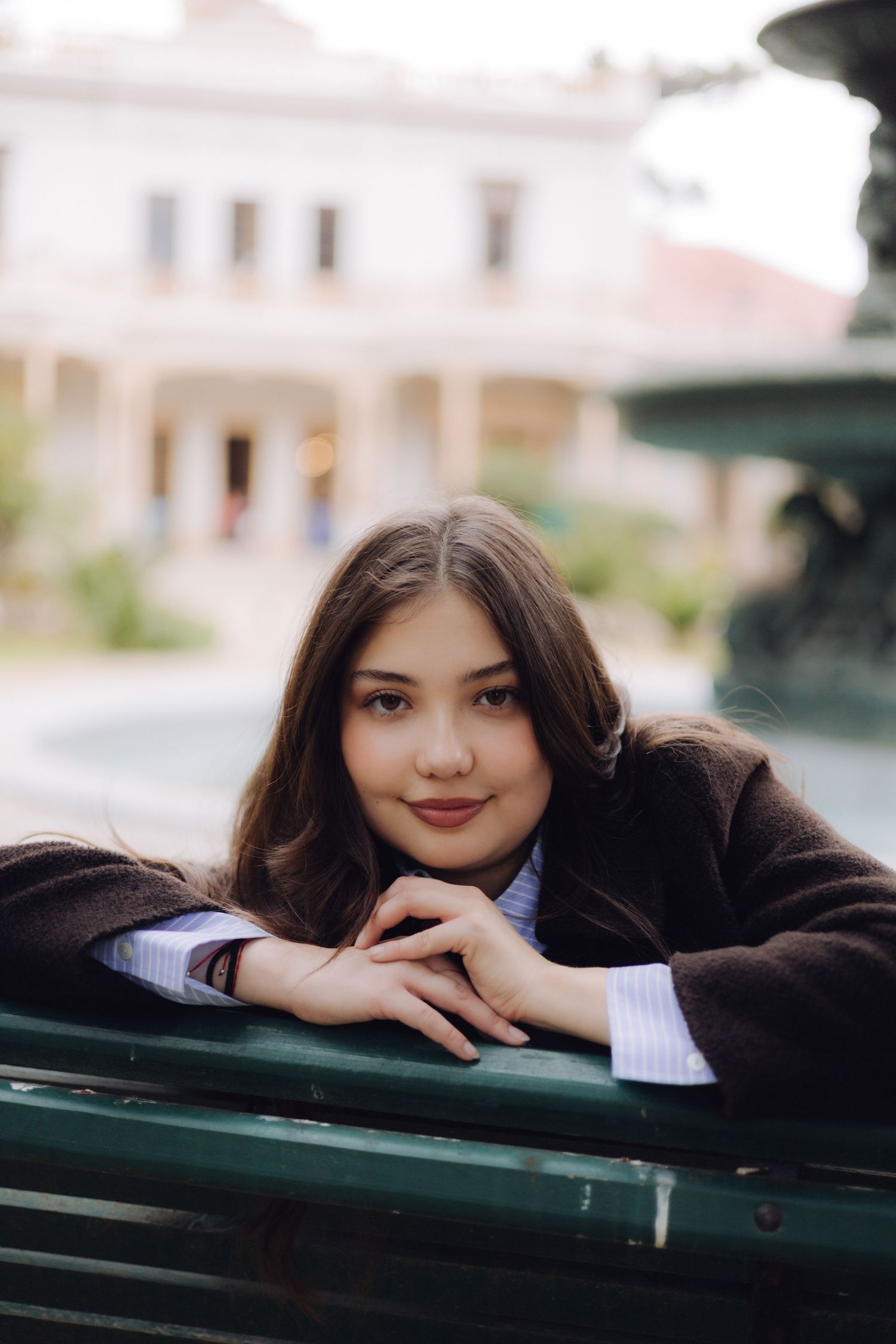 Young woman with long brown hair leaning on a green bench, smiling softly in front of a fountain.