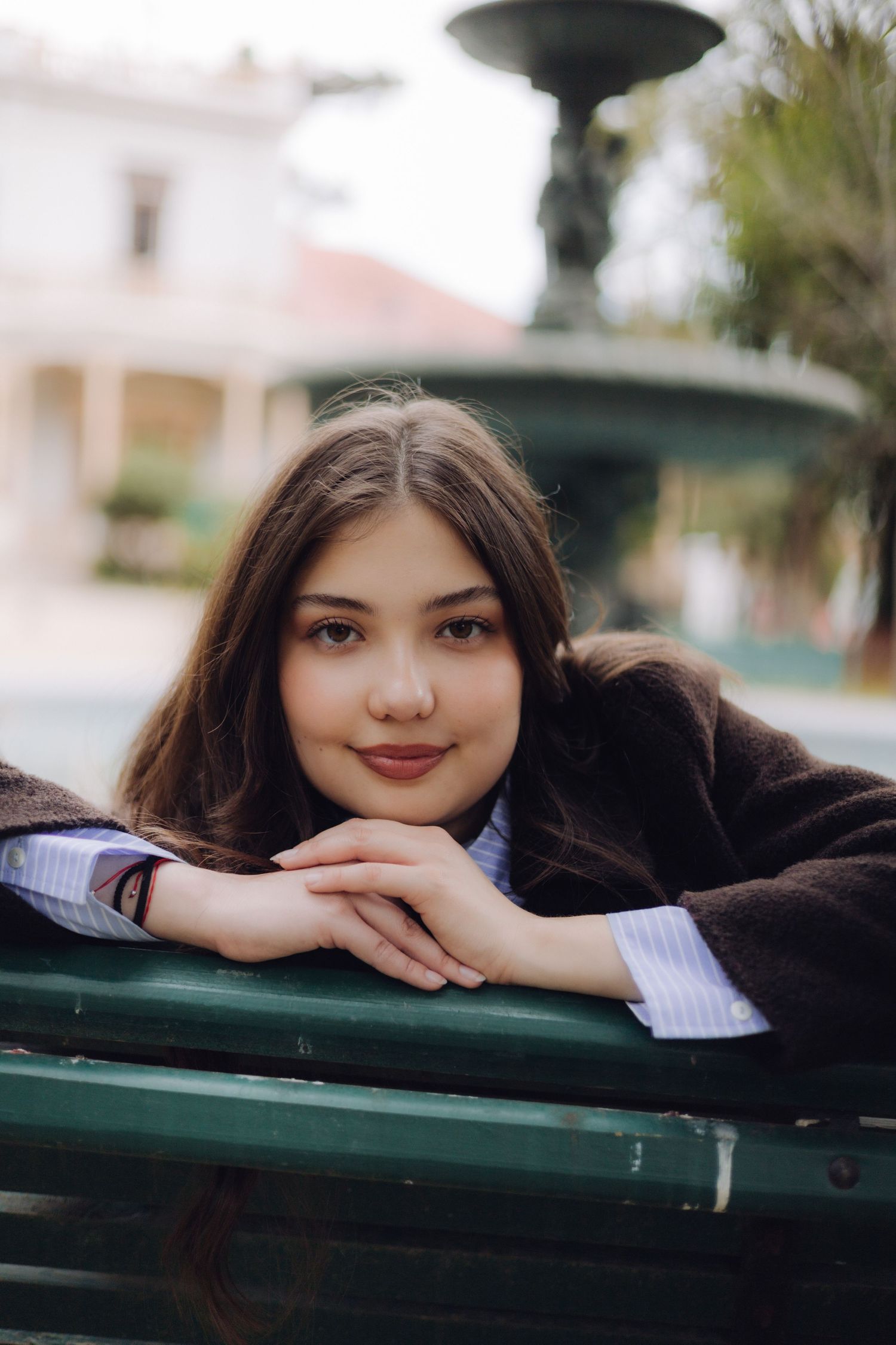 Young woman with long brown hair resting on a green bench near a fountain in a town square.