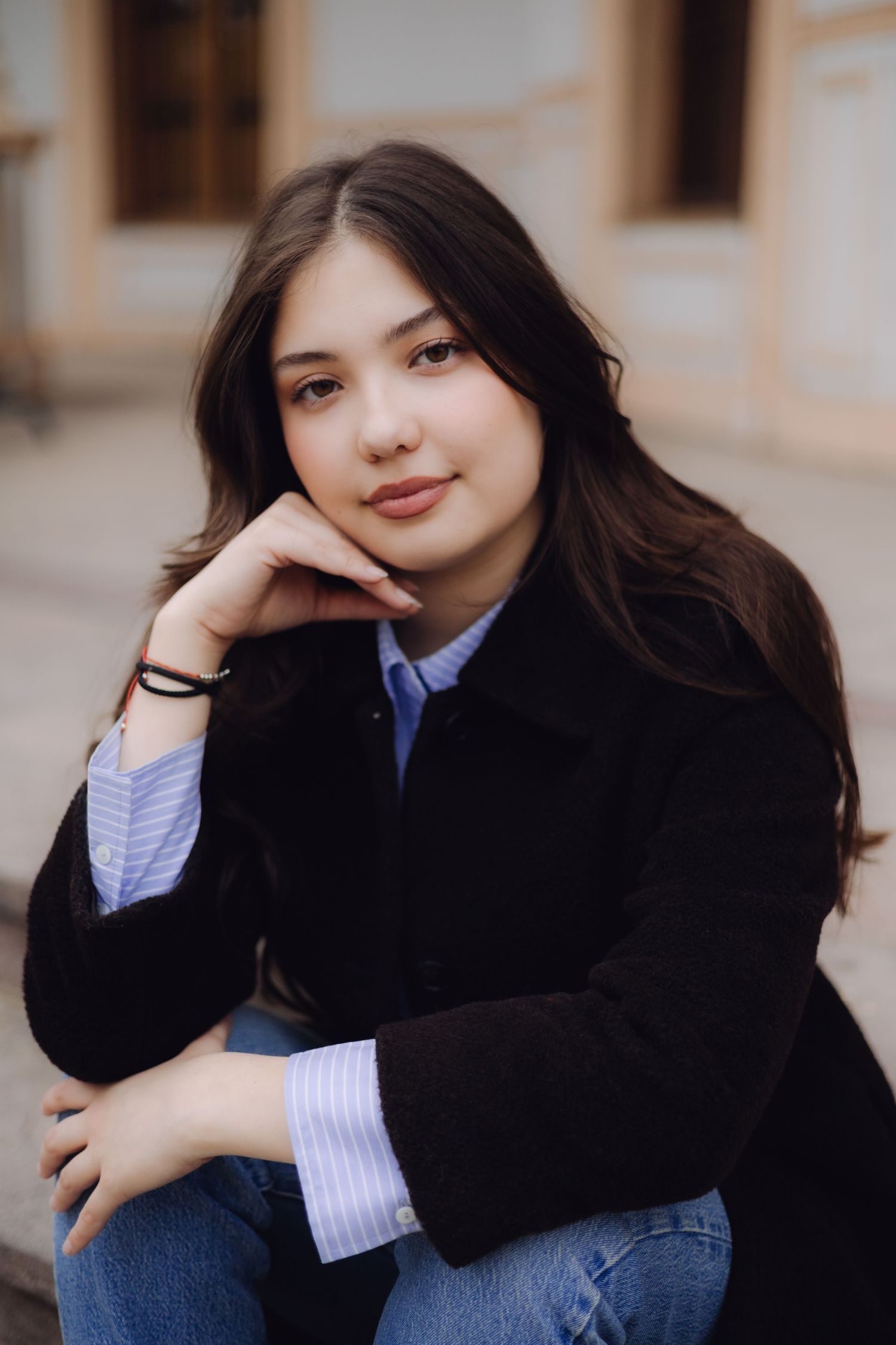 Young woman with dark hair in black blazer and jeans sits outdoors, resting chin on hand, smiling softly.