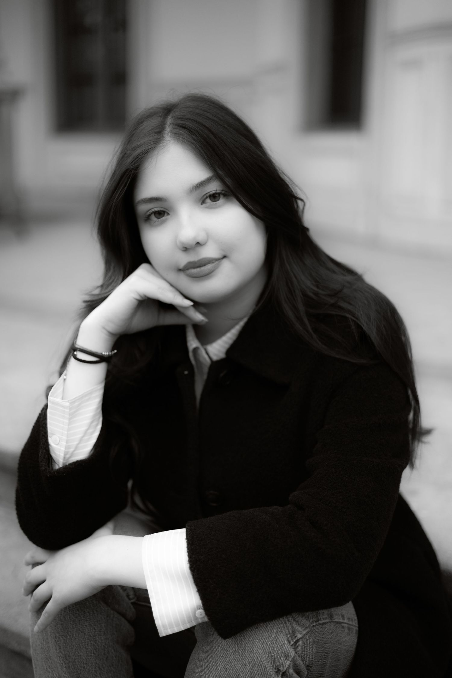 Black and white portrait of a young woman with long dark hair wearing a black coat, resting chin on hand.