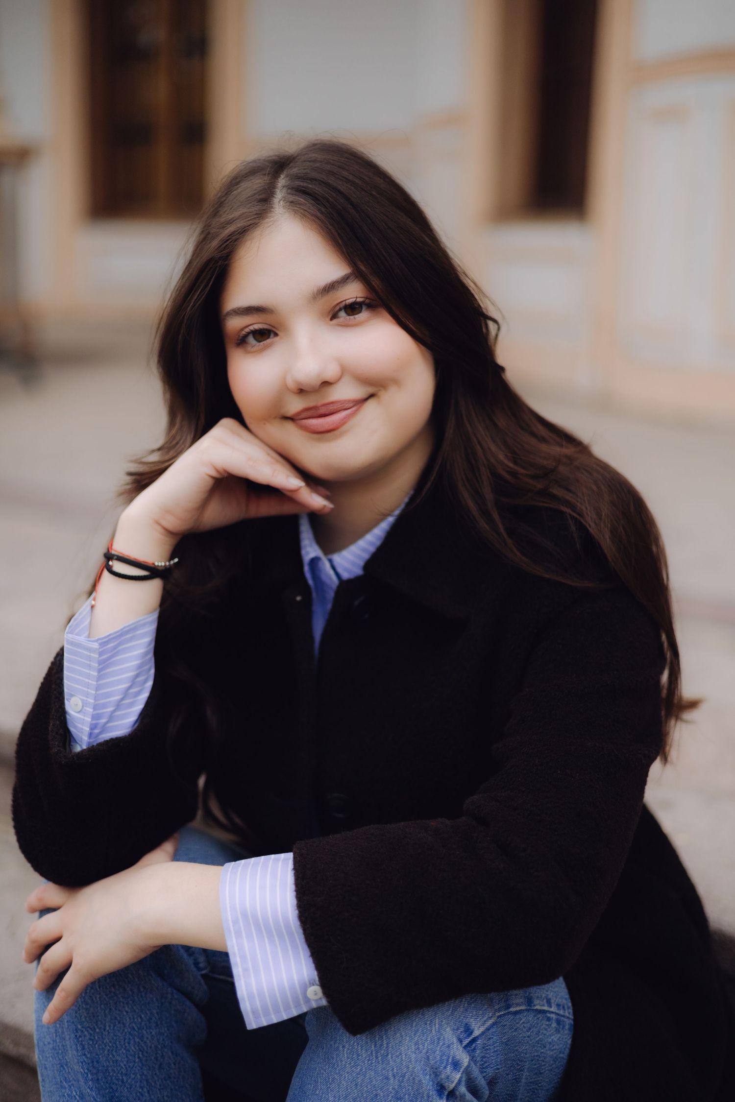 Young woman with long dark hair smiling, wearing a black coat and blue striped shirt, sitting outdoors.