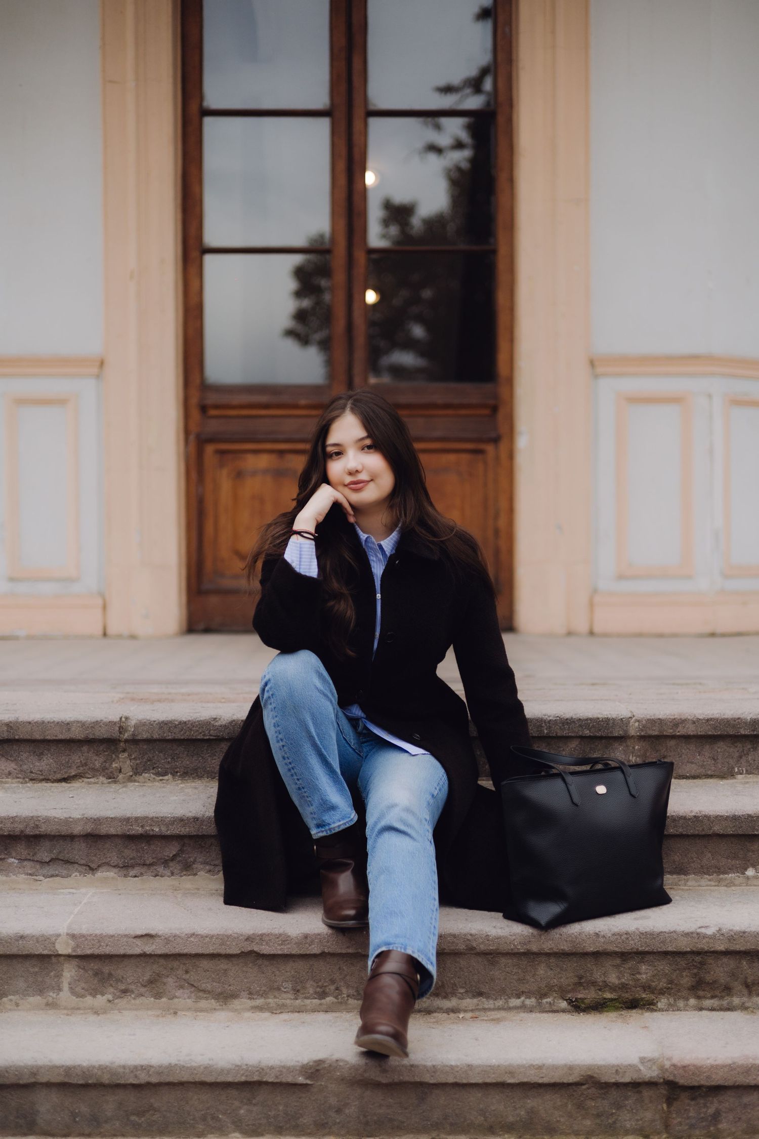Young woman in black coat and jeans sitting on stone steps with a black bag, posing thoughtfully outdoors.
