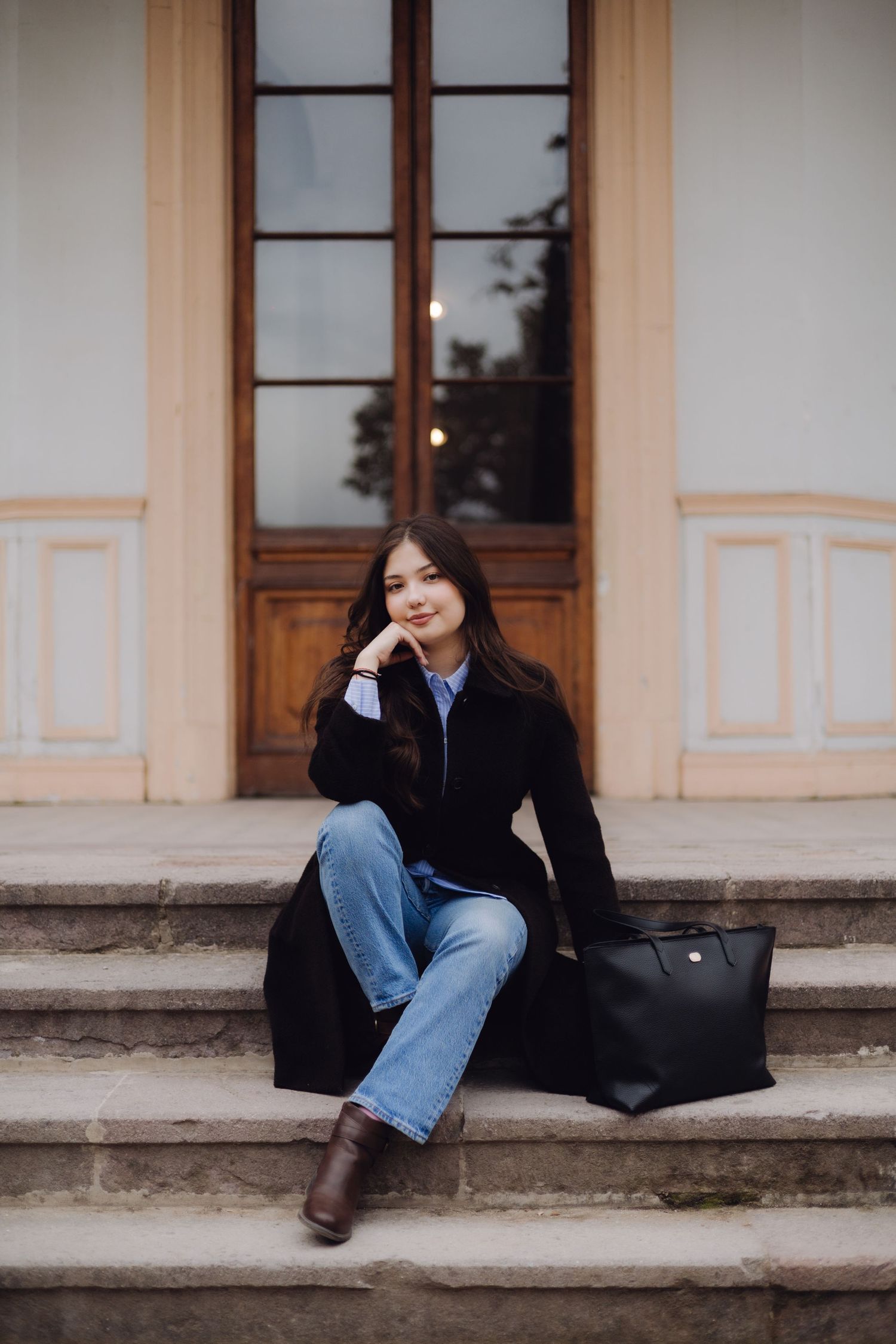 Young woman in black coat and jeans sits on stone steps with black tote bag, smiling softly outdoors.